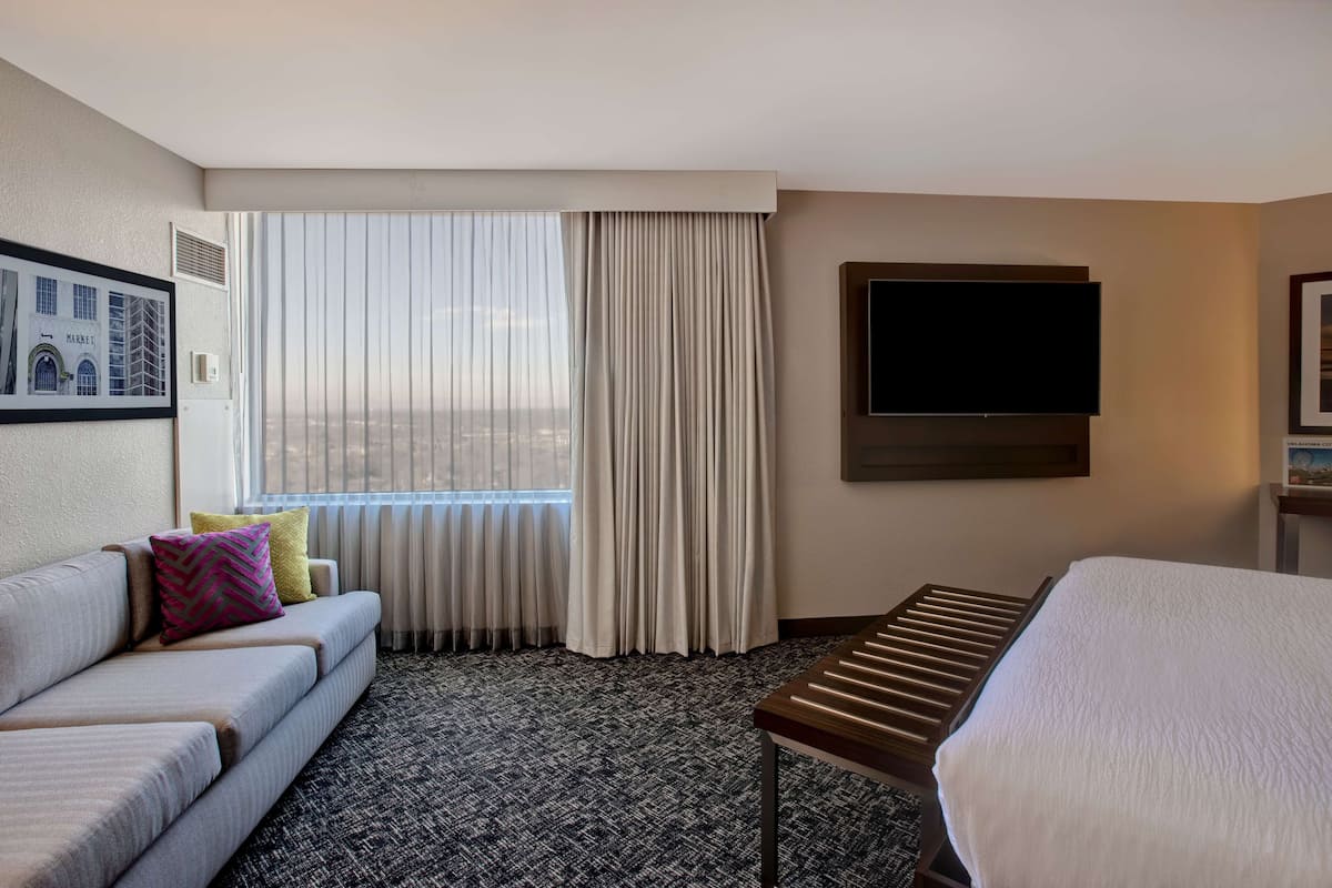 A hotel room with a sofa, a bed, a TV, and a window with curtains.