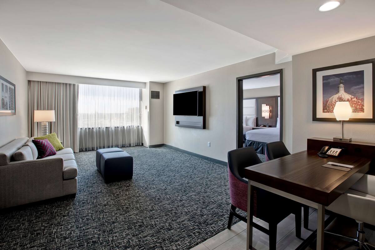 A modern hotel room with a sofa, a small table, a desk, a flat-screen TV, a lamp, and a framed picture on the wall.
