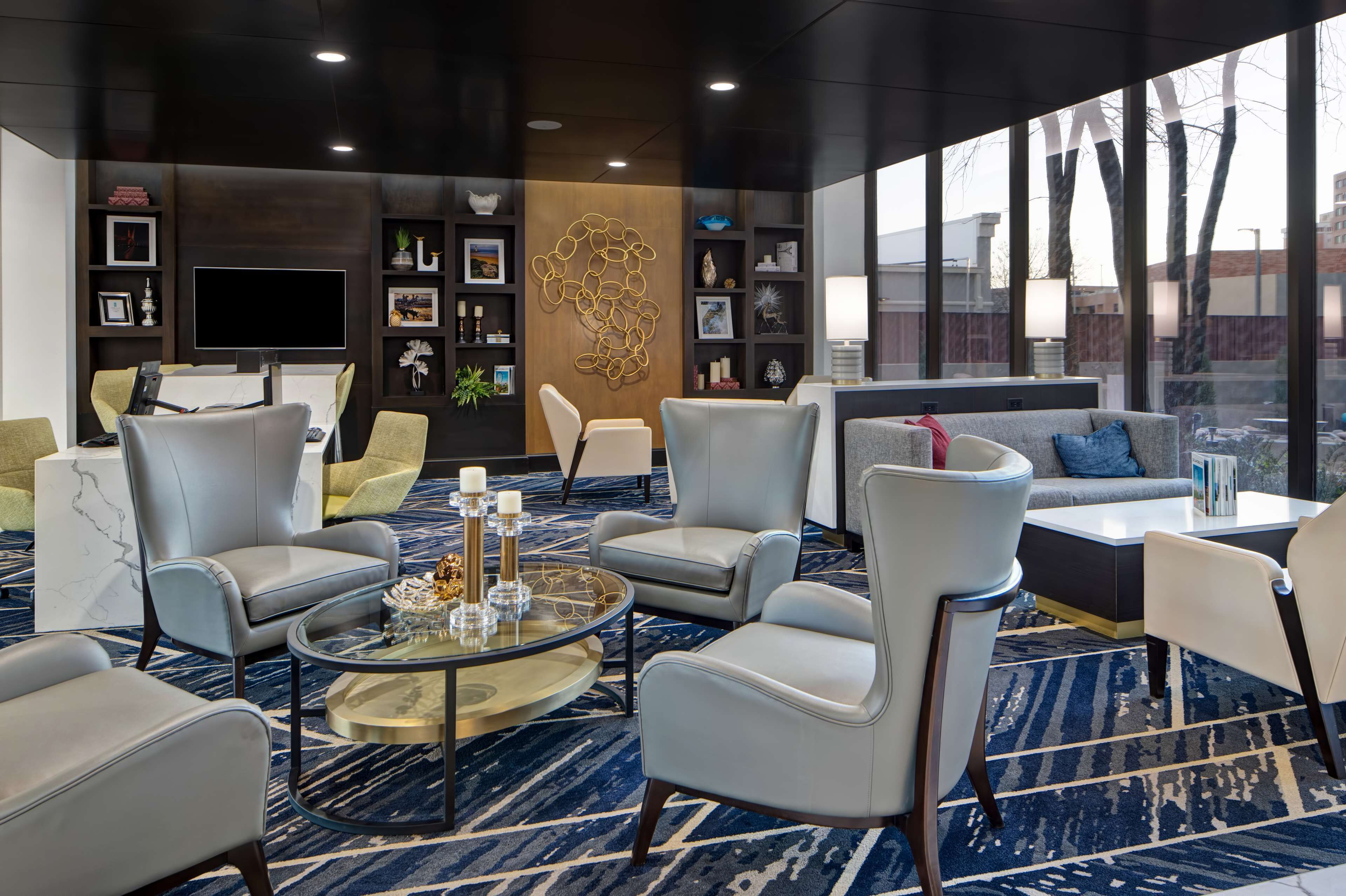 A modern lounge area with leather chairs, a coffee table, and a large TV screen. There are shelves with decorative items and a large window offering a view of the outside.