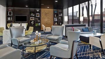 A modern lounge area with leather chairs, a coffee table, and a large TV screen. There are shelves with decorative items and a large window offering a view of the outside.