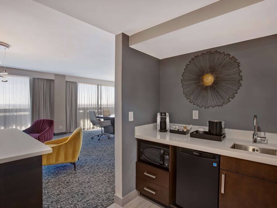 Una habitación de hotel moderna con kitchenette, un gran elemento decorativo en la pared y vistas de la ciudad a través de amplios ventanales.