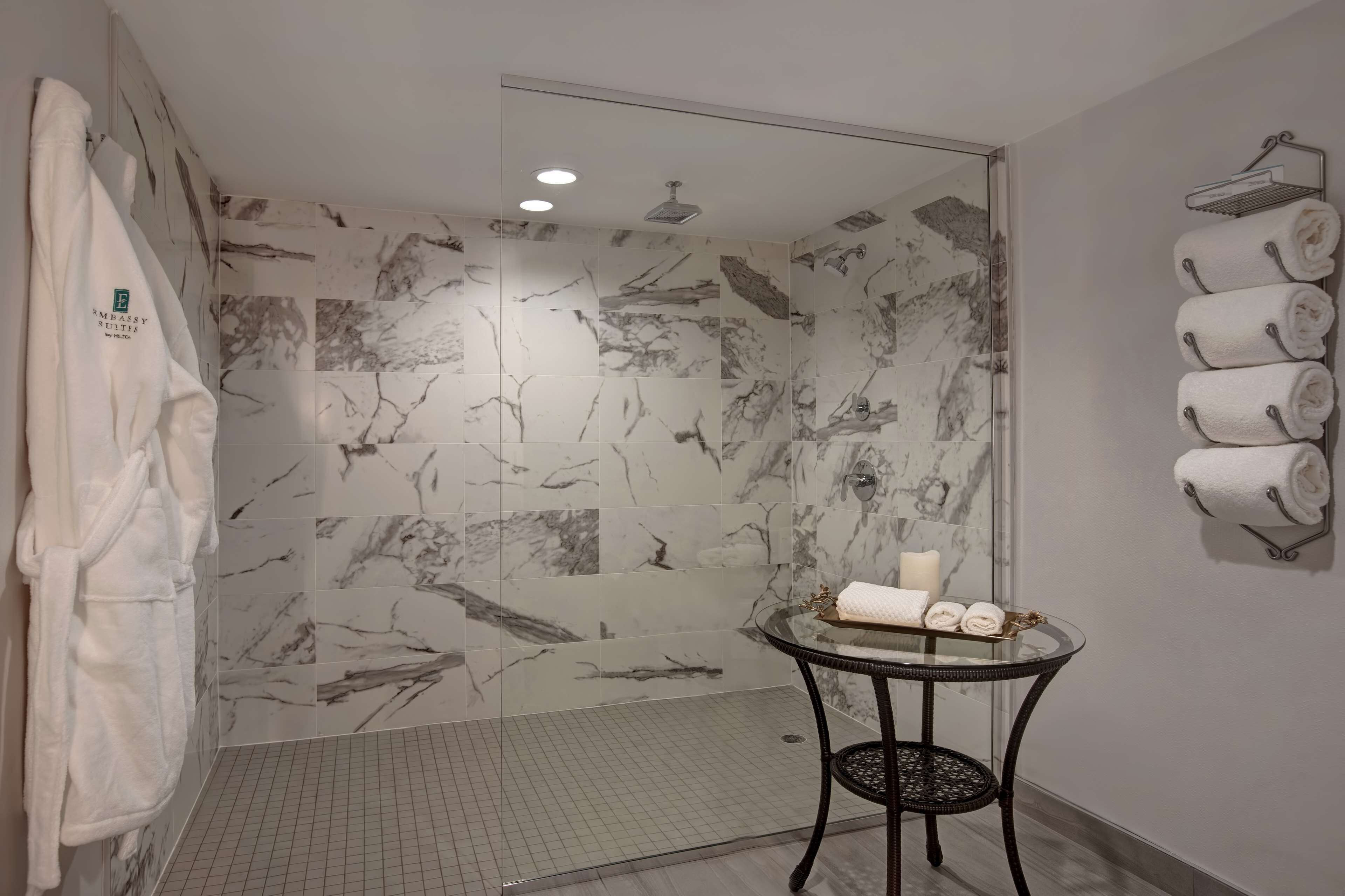 Une salle de bain moderne avec des carreaux de marbre, une douche en verre, une table en verre avec des serviettes et un peignoir accroché au mur.