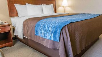 A hotel room with a wooden headboard, a bed with white and brown bedding, a blue blanket, a nightstand with a phone, and a lamp.
