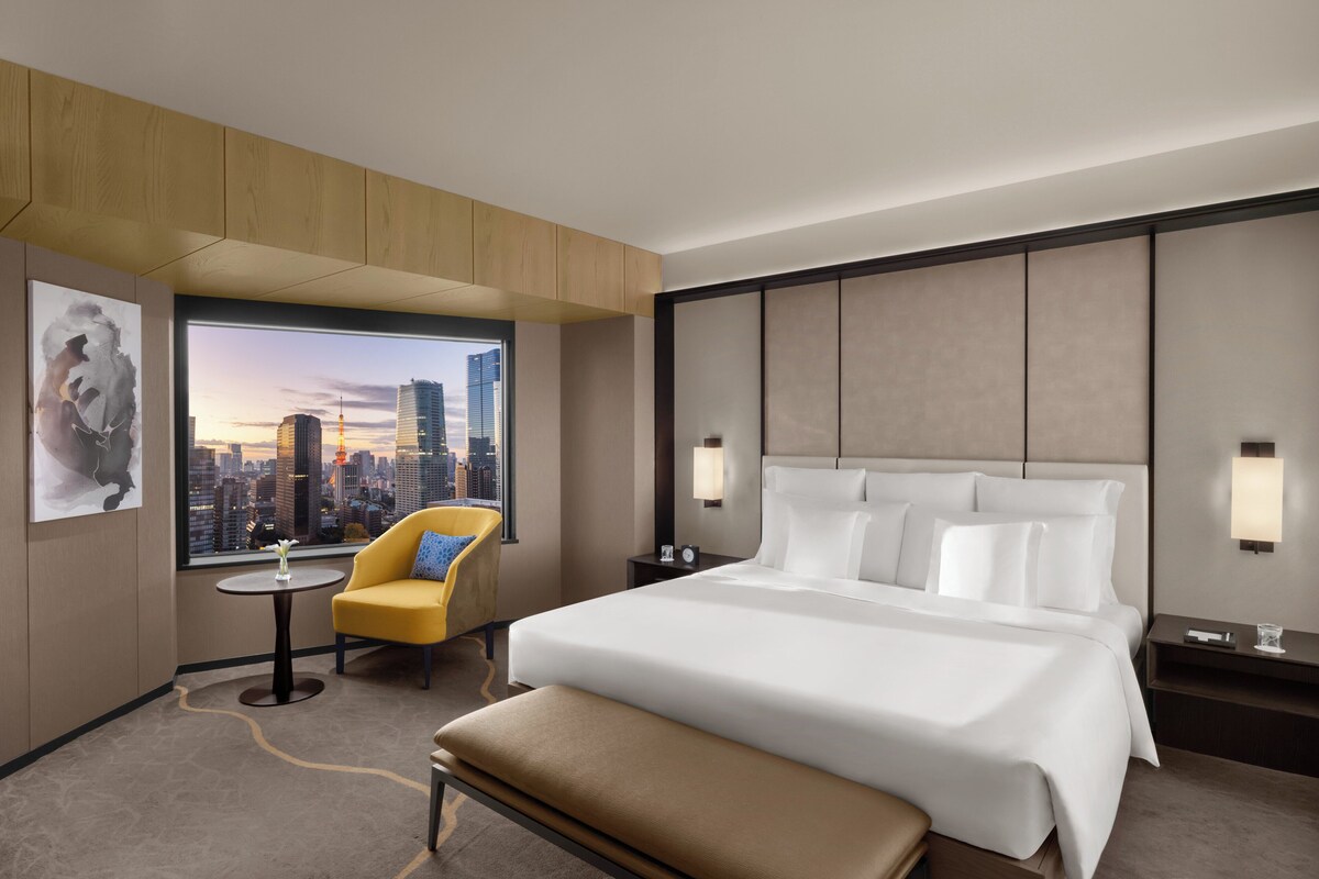 A hotel room with a large bed, a small table, a yellow armchair, and a view of a cityscape.