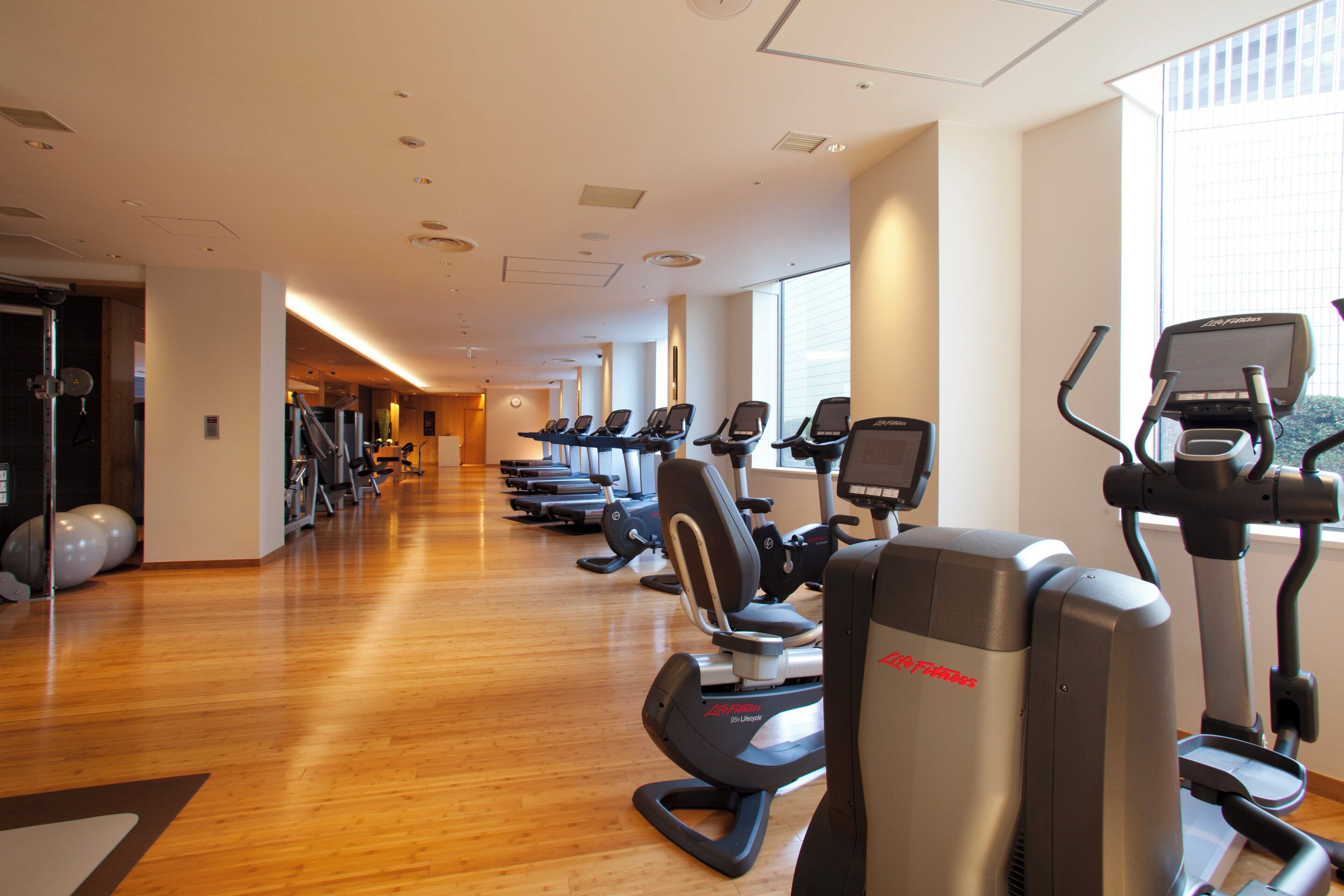 A gym with multiple cardio machines and a wooden floor.