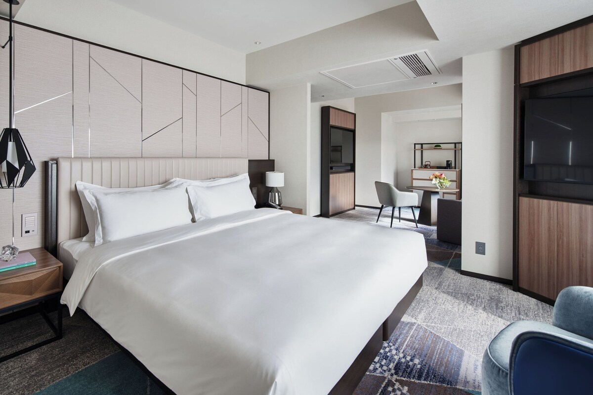 A modern hotel room with a large bed, a desk with a chair, a TV, and a bathroom visible through an open door.