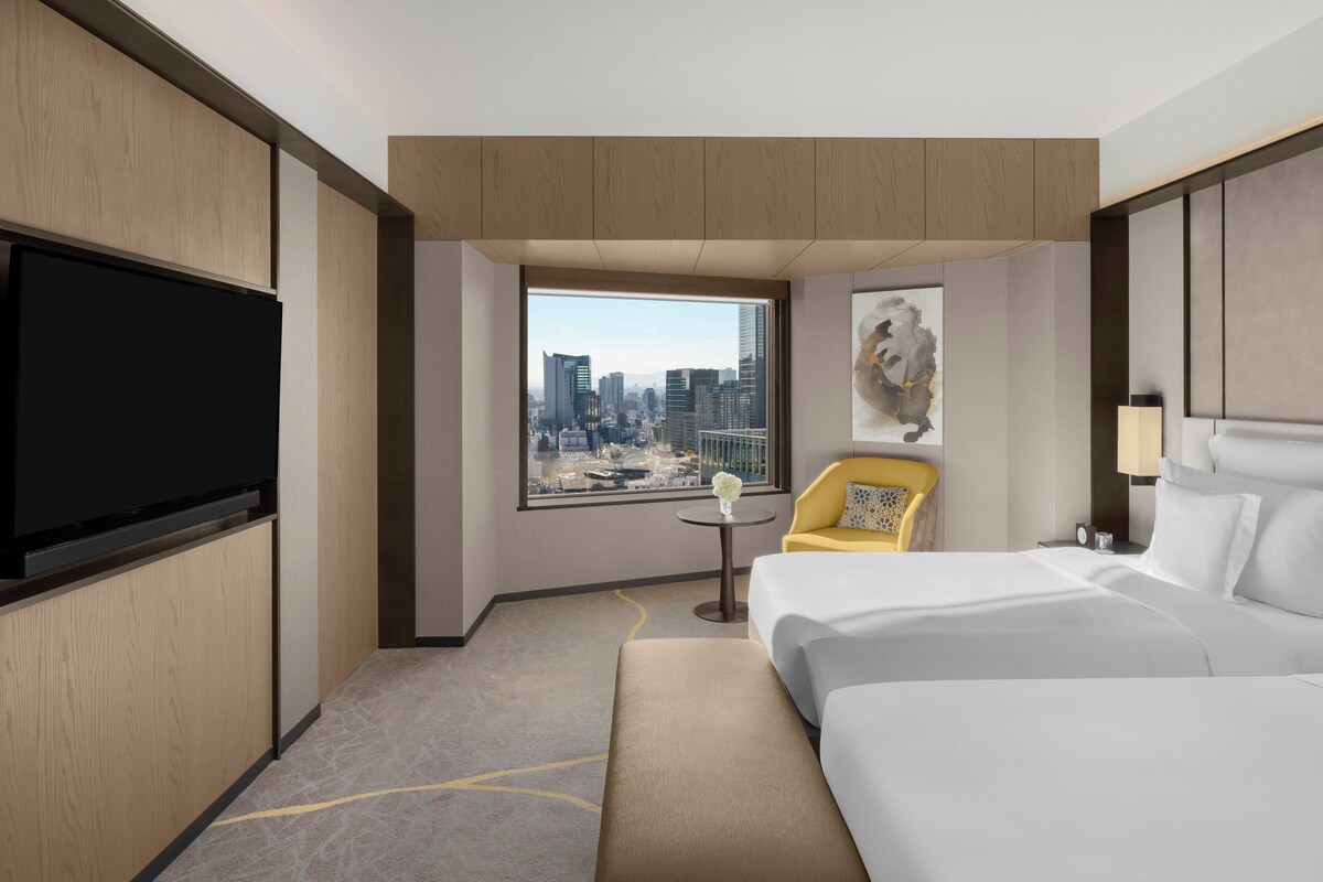 A hotel room with a large bed, a TV, a chair, a small table, and a window with a city view.