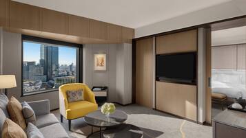 Habitación de hotel moderna con un ventanal grande que ofrece vistas a la ciudad, un sofá, un sillón amarillo, un televisor de pantalla plana y una cama con sábanas blancas.