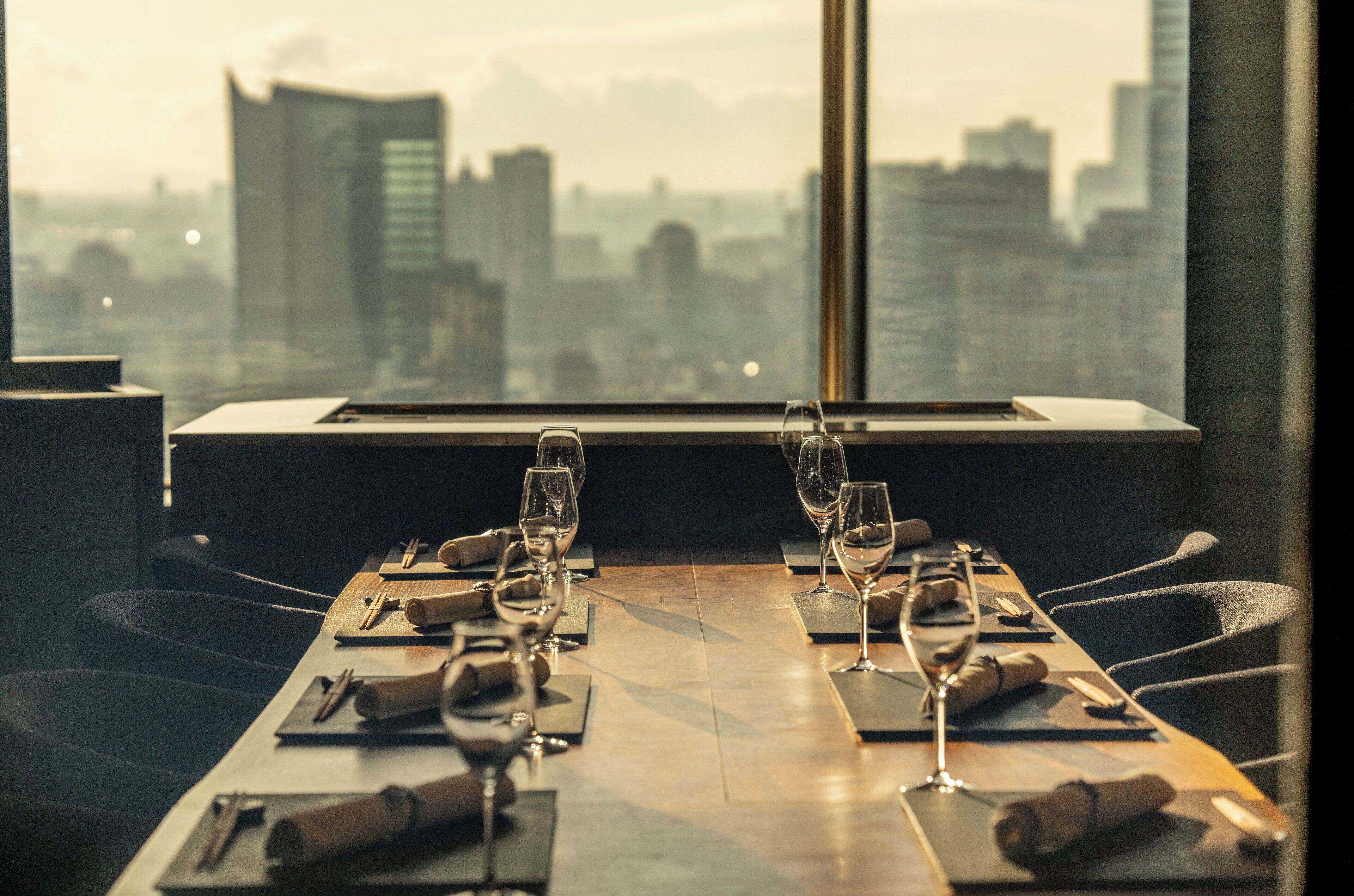 A dining table set for four with wine glasses and cutlery, overlooking a cityscape through a large window.