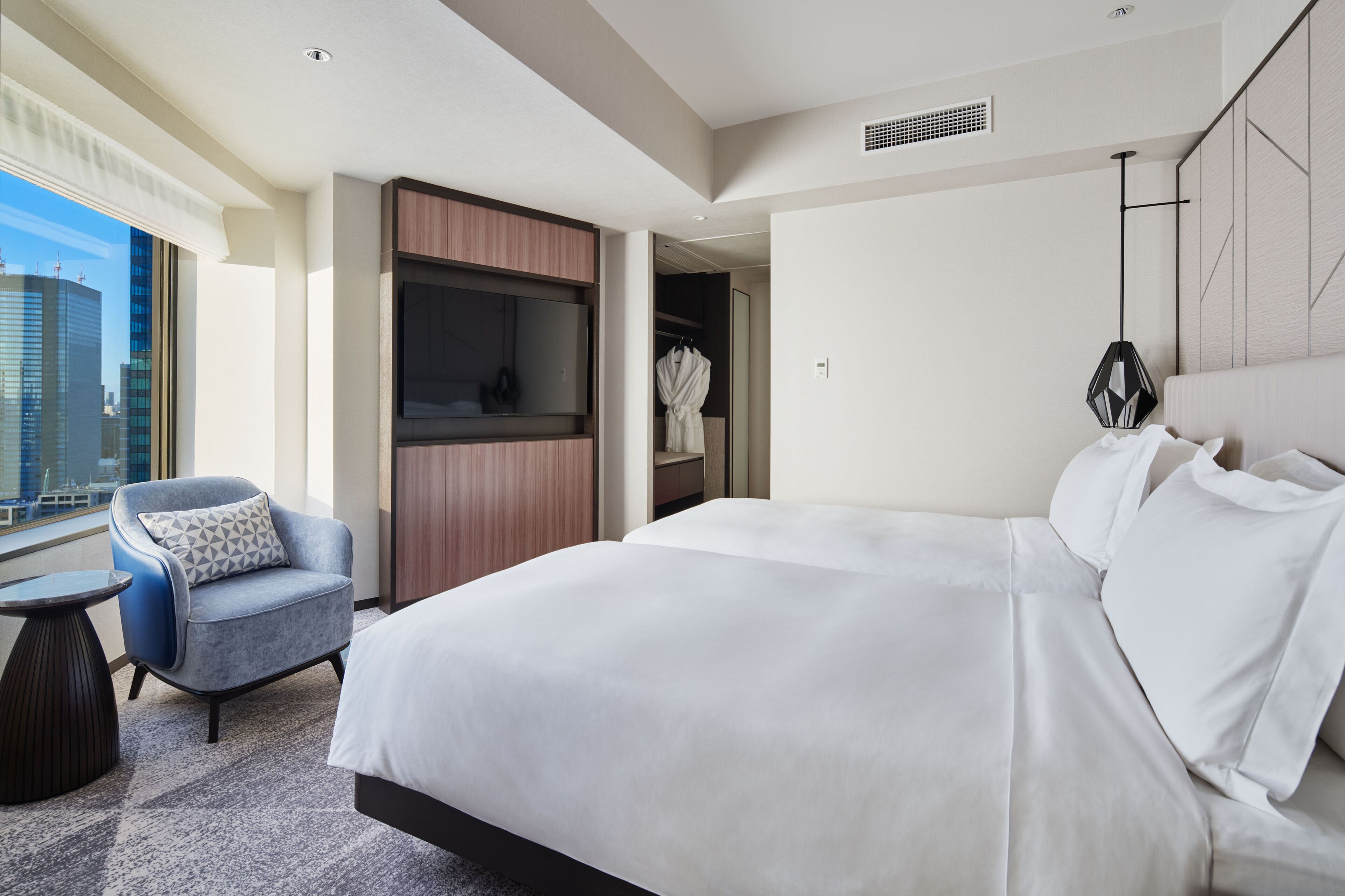 A modern hotel room with a large bed, a seating area with a chair, a wardrobe, and a view of the cityscape.