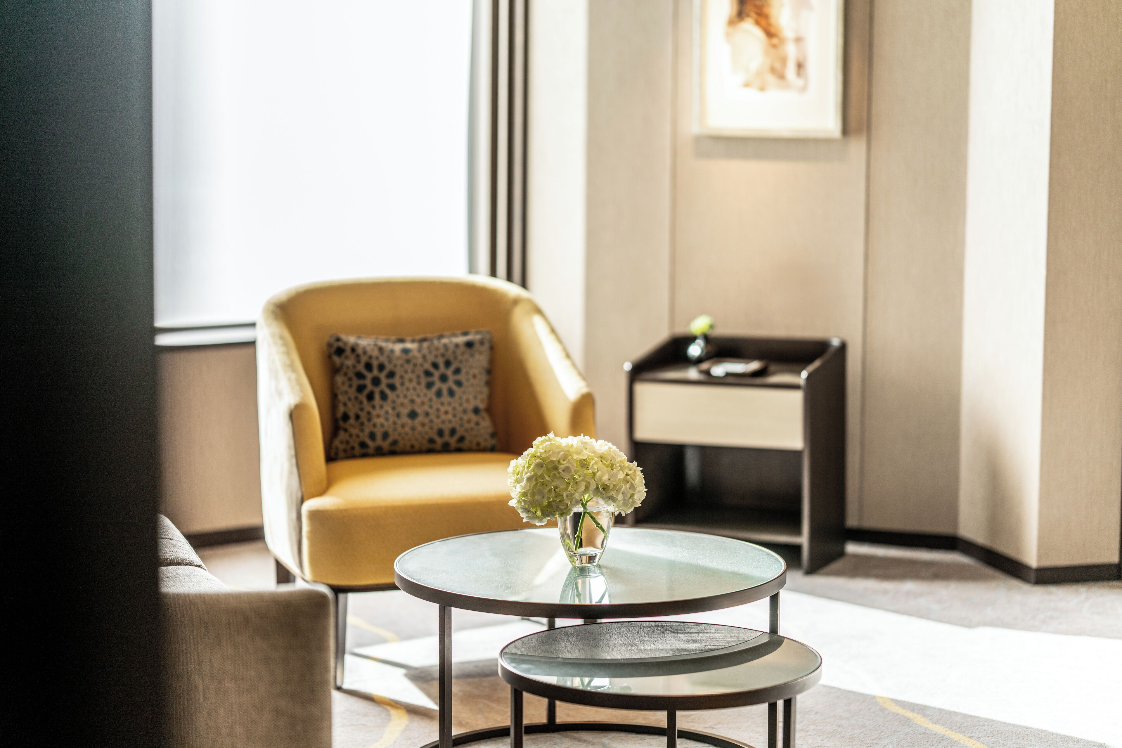 A modern room with a yellow armchair, a small round table with a vase of flowers, and a side table with a plant.