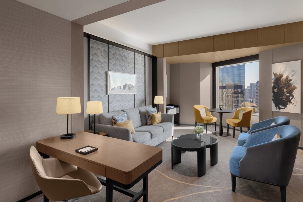 A modern hotel room with a sofa, armchairs, a dining table, and a city view.