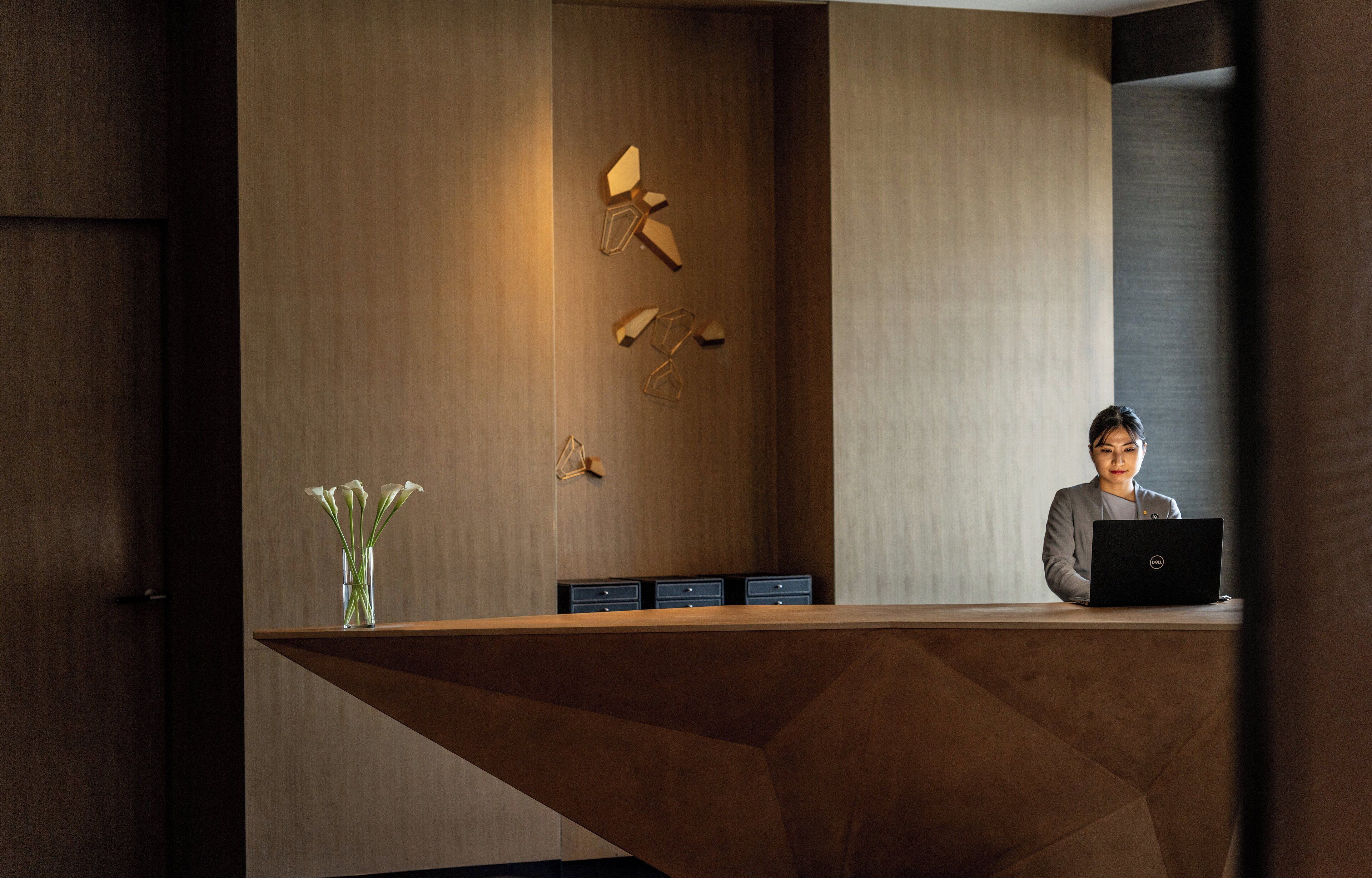 A modern reception desk with a wooden finish, a vase with white flowers, and a laptop on the desk.
