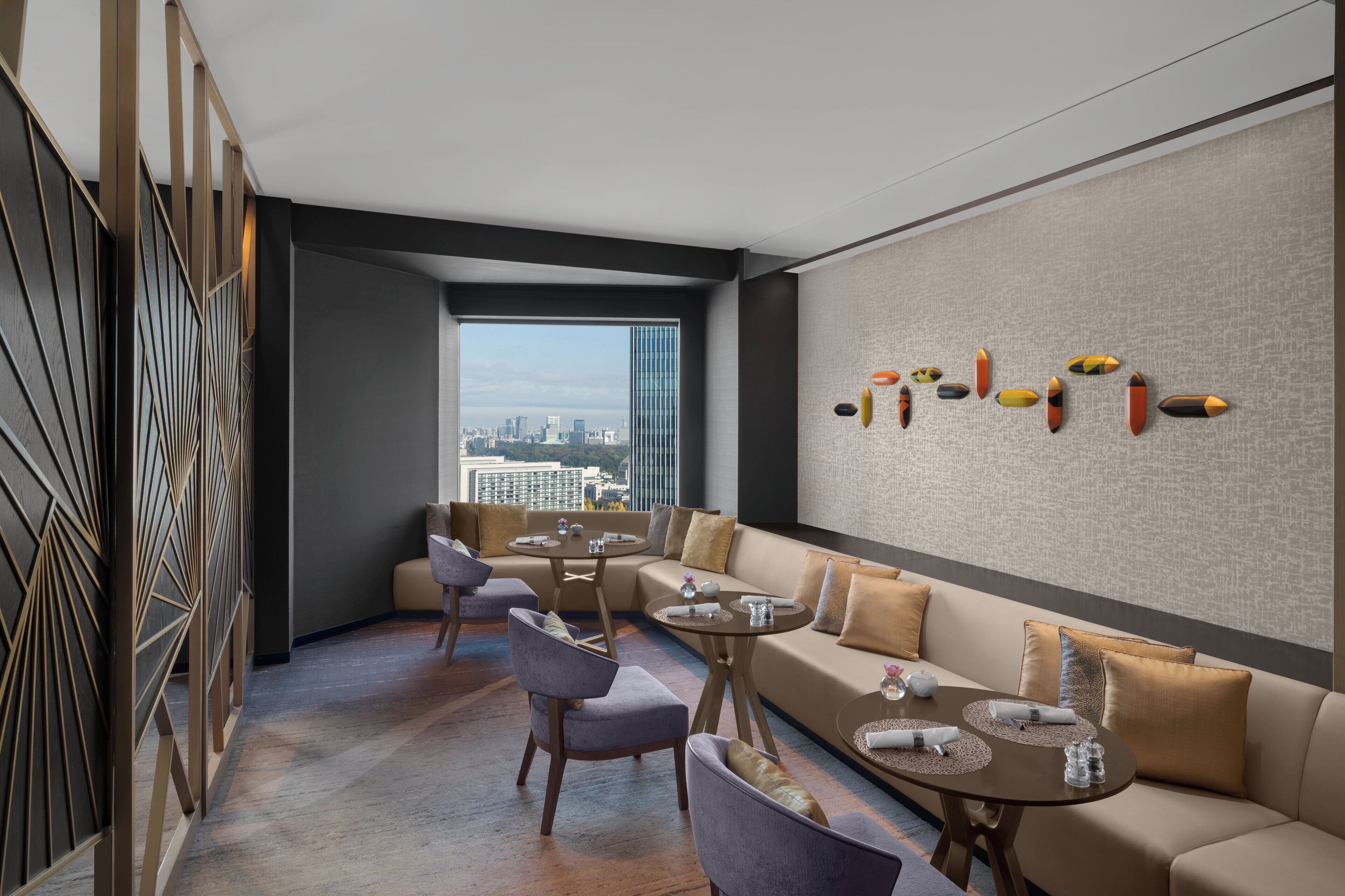 A modern lounge area with a sofa, chairs, and a dining table, featuring a city view through large windows.