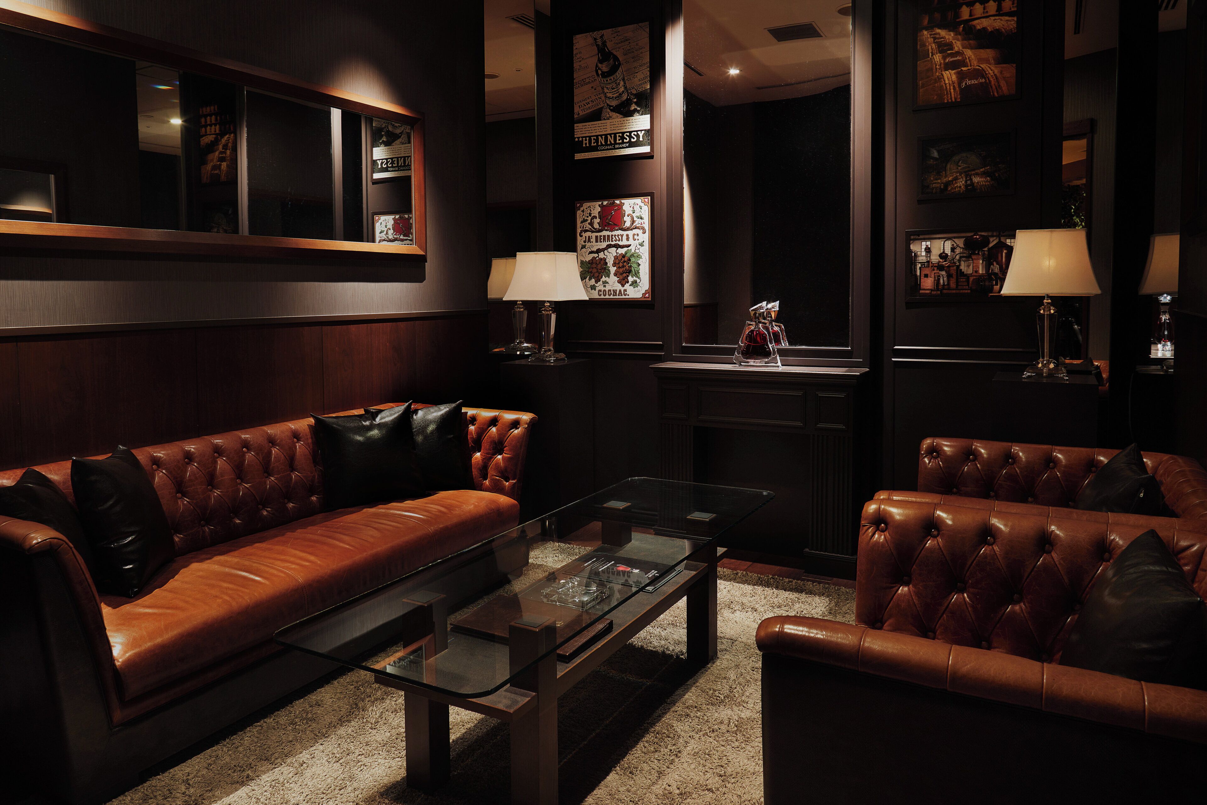 A dark-themed living room with leather sofas, a glass coffee table, and wall-mounted lamps.