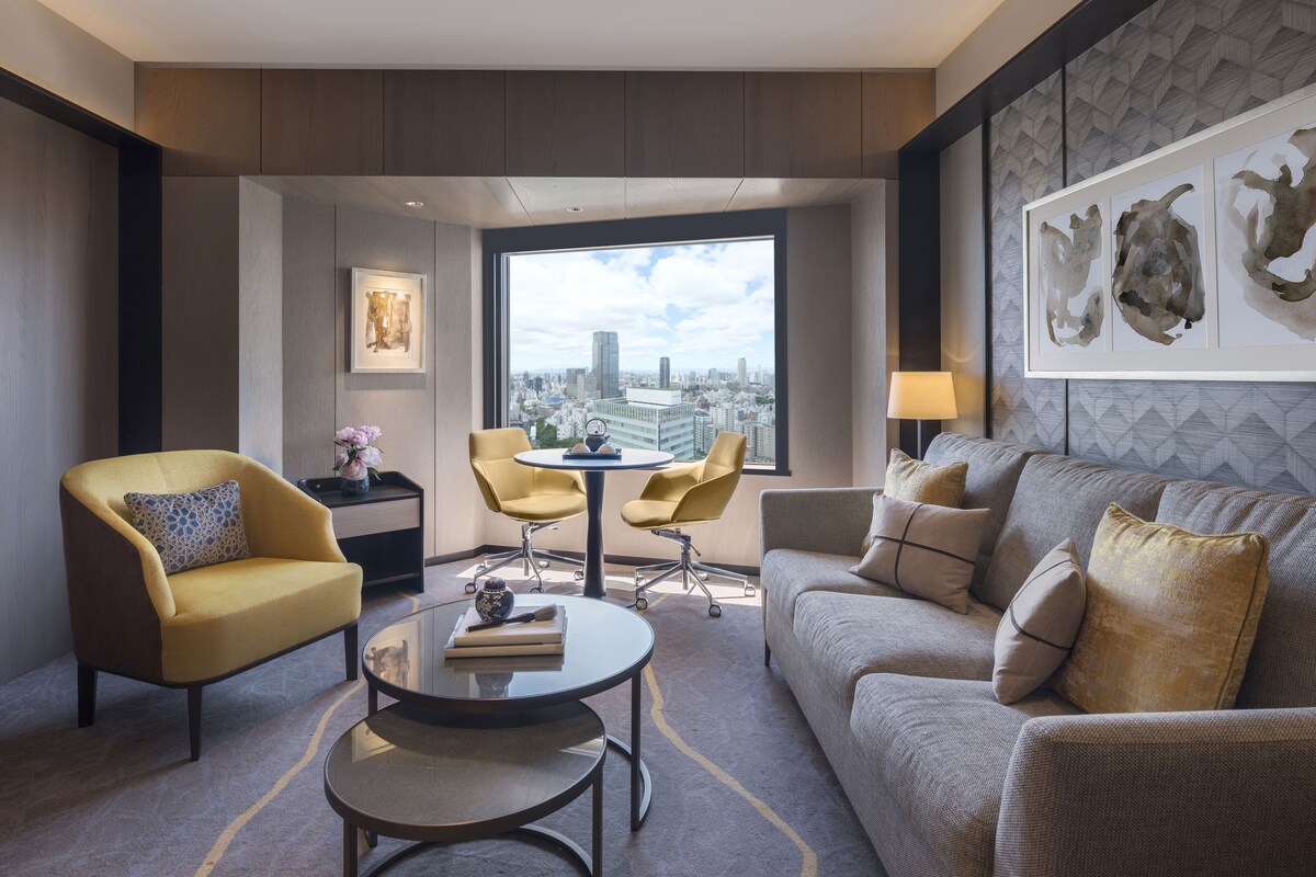 A modern hotel room with a large window offering a city view, a sofa, a yellow armchair, a round coffee table, and a small desk with a lamp.