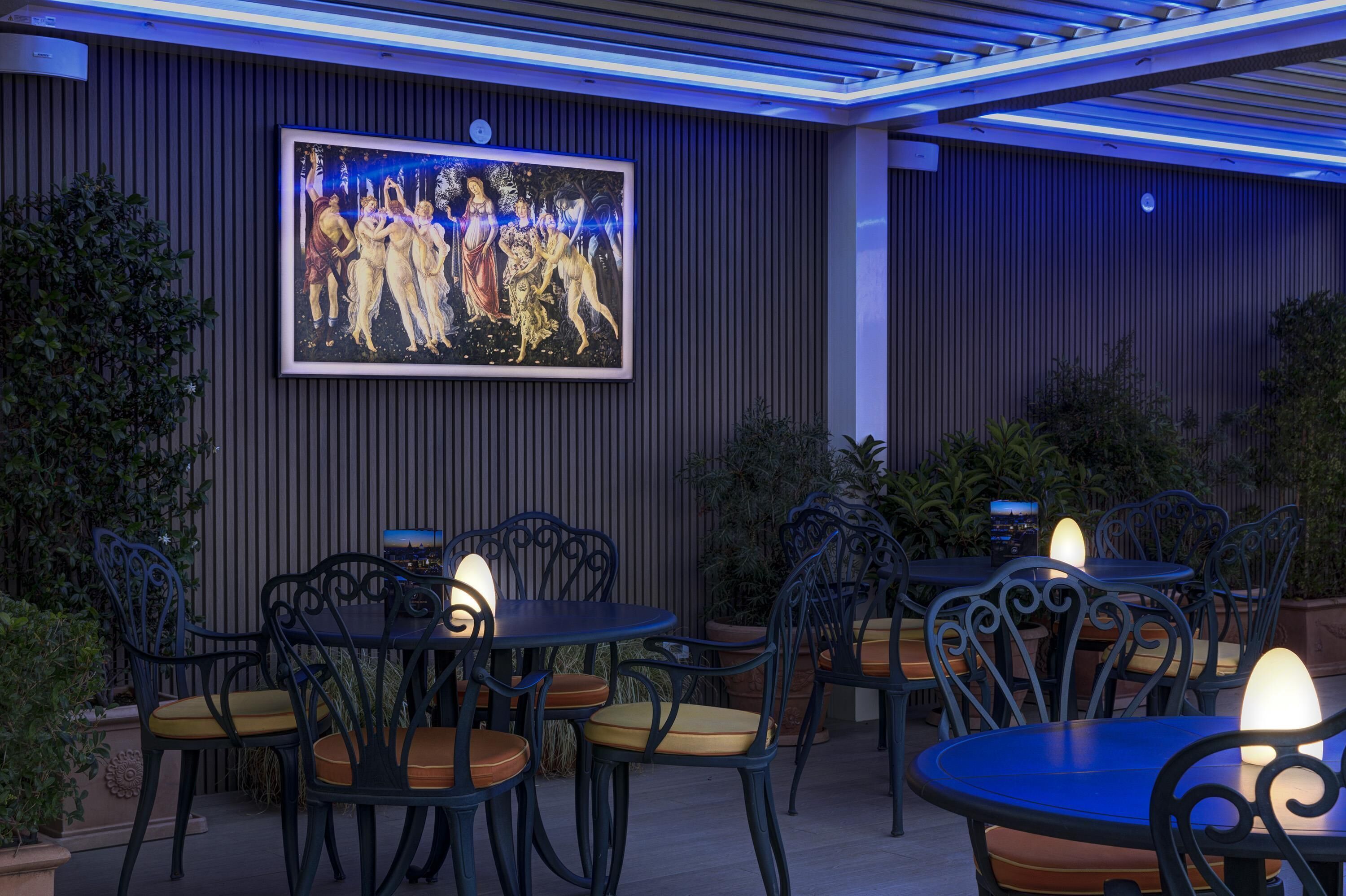 An outdoor patio area with tables and chairs, a large framed painting on the wall, and ambient lighting.