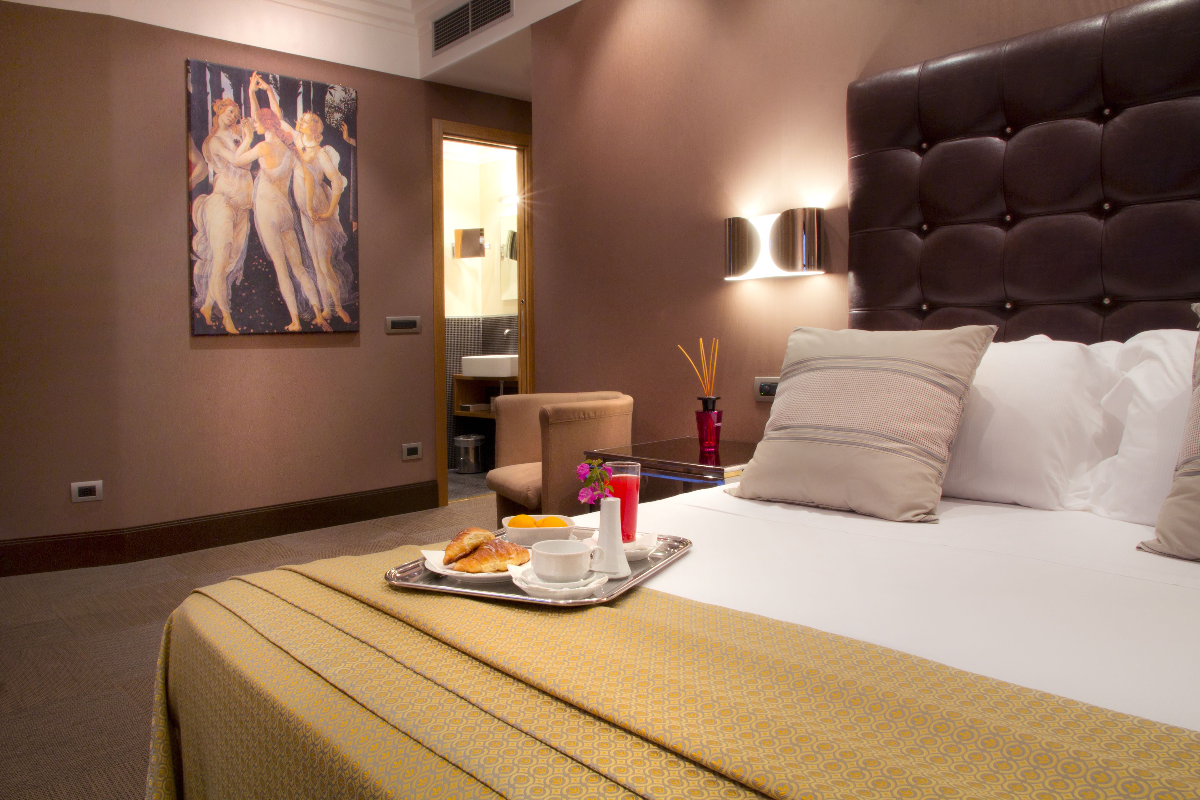 A hotel room with a large bed, a painting on the wall, a tray with breakfast items, and a bathroom visible through an open door.