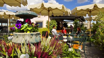 An outdoor cafe with tables and chairs under umbrellas, surrounded by various plants and flowers.