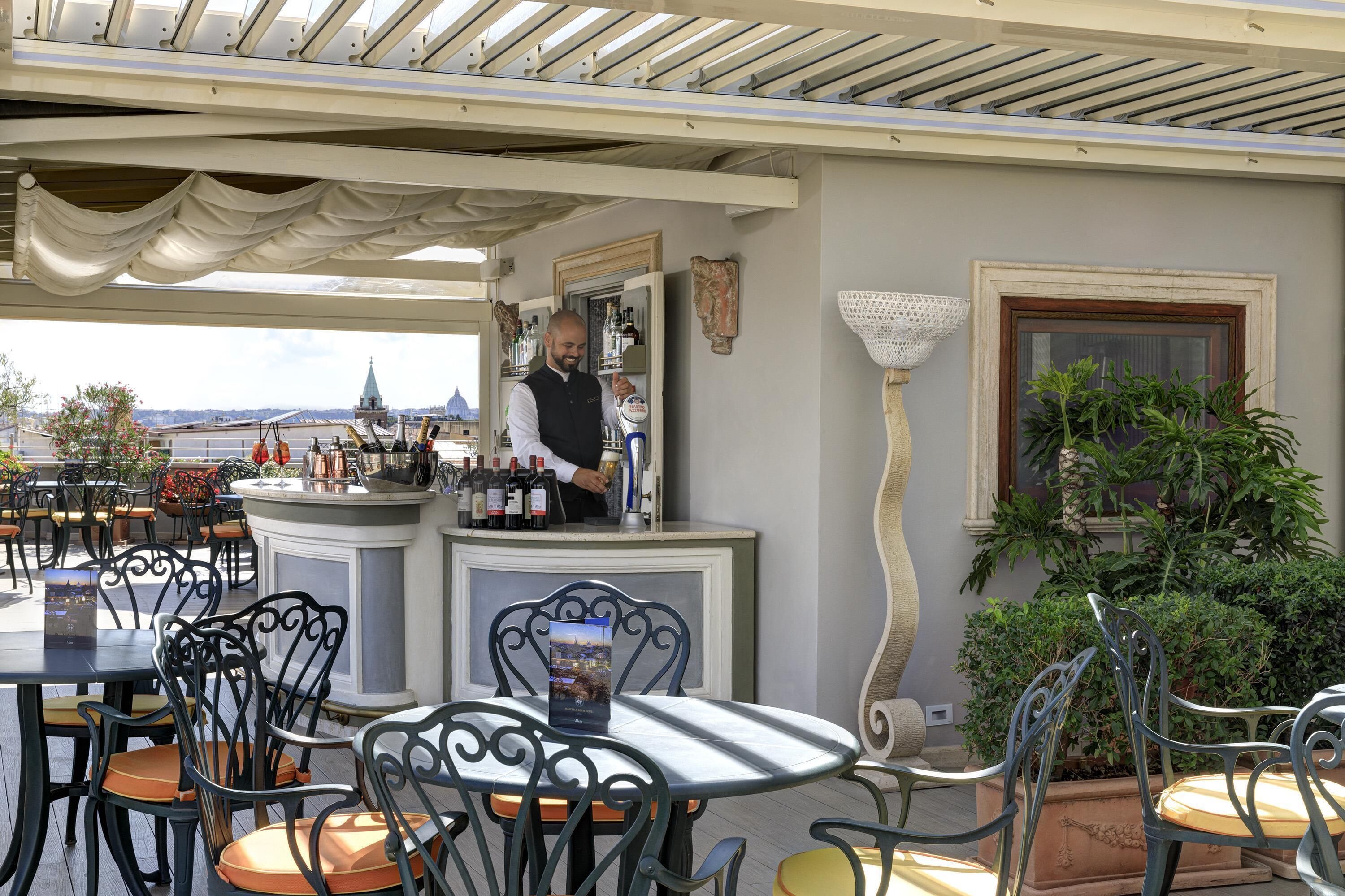 An outdoor bar area with a bartender, tables, chairs, and a view of a cityscape.