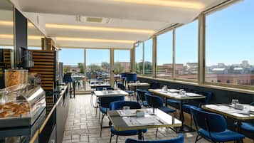 A restaurant with tables and chairs, a counter with food items, and large windows offering a city view.
