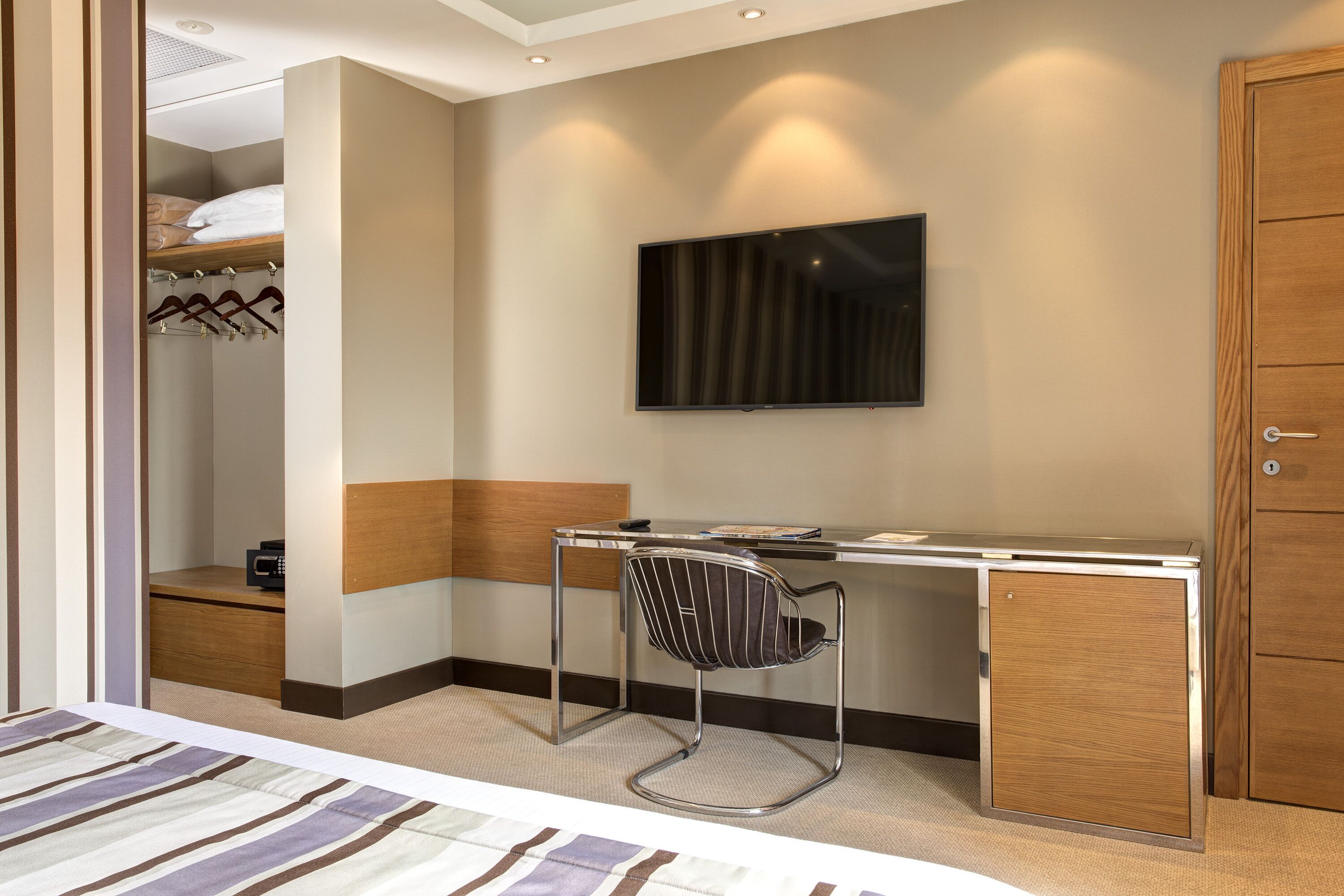A hotel room with a bed, a desk with a chair, a TV mounted on the wall, and a wardrobe.