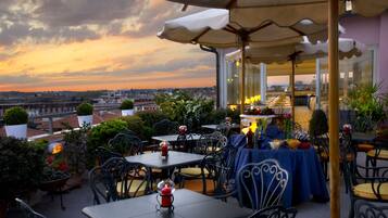 An outdoor patio with tables and chairs, a view of the cityscape, and a sunset in the background.