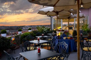 An outdoor patio with tables and chairs, a view of the cityscape, and a sunset in the background.
