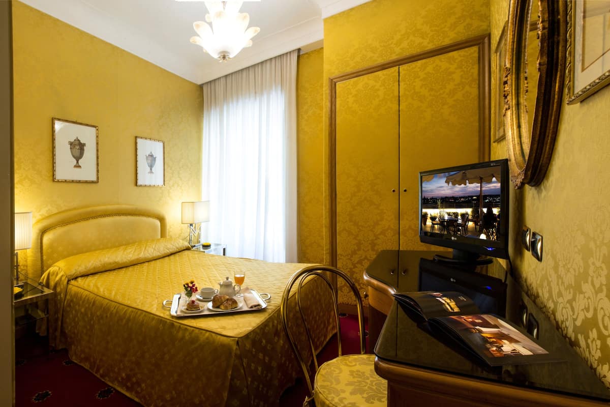 Camera d'albergo con un letto, una scrivania con un computer, un vassoio con prodotti per la colazione e una televisione che mostra una vista sulla città.