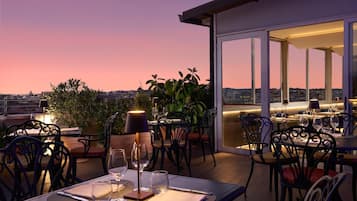 En tagterrasse med borde og stole, plantekasser og udsigt over byens skyline i skumringen.