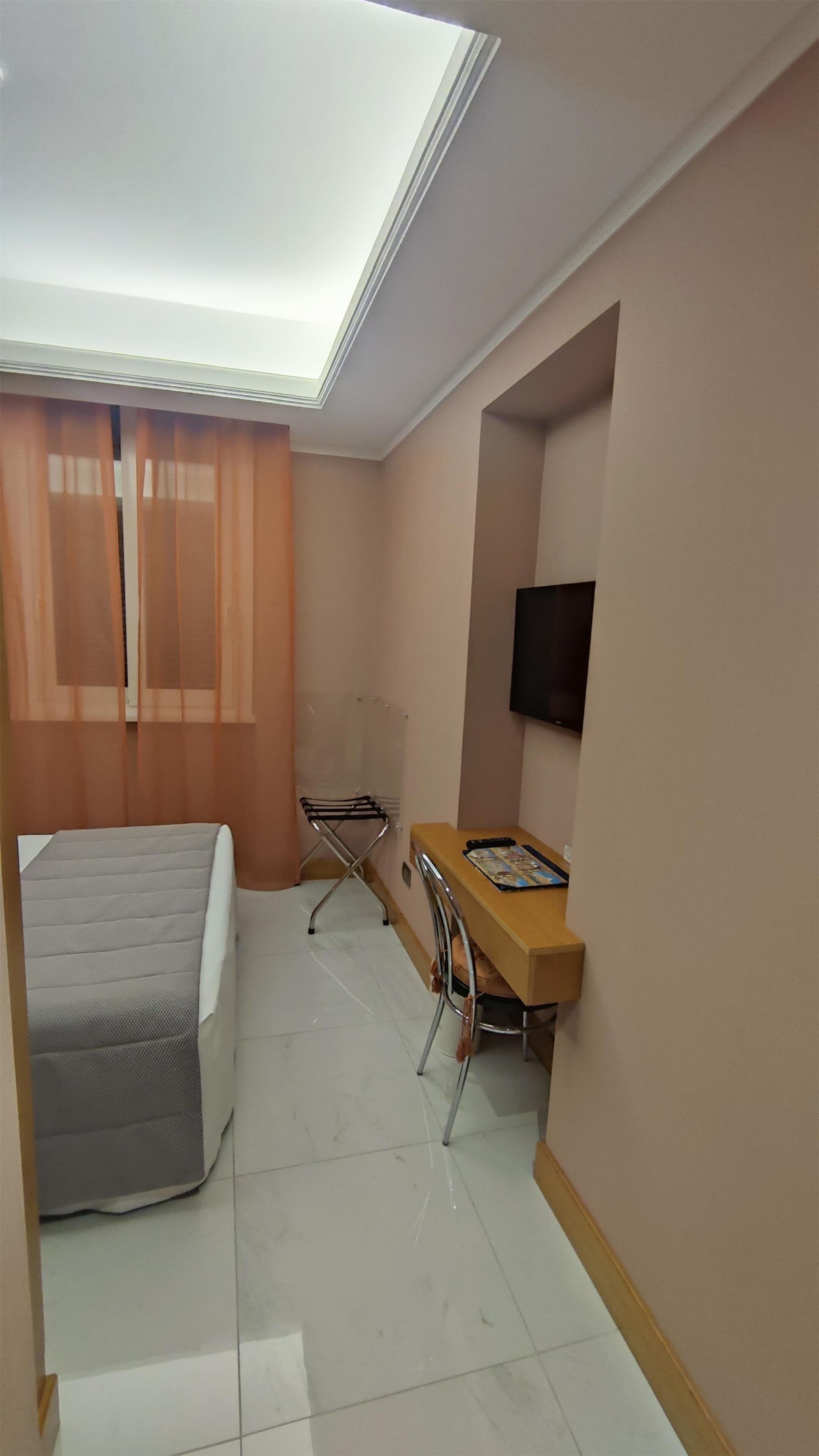 A hotel room with a bed, a desk with a chair, a TV mounted on the wall, and a window with curtains.