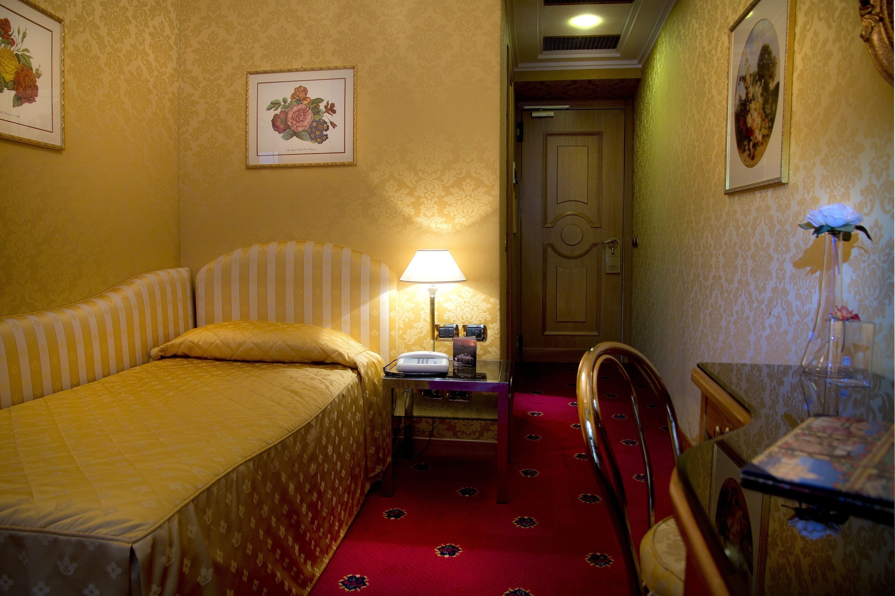 Et hotelværelse med en enkelt seng, et natbord med lampe, en stol og et lille bord med en vase.