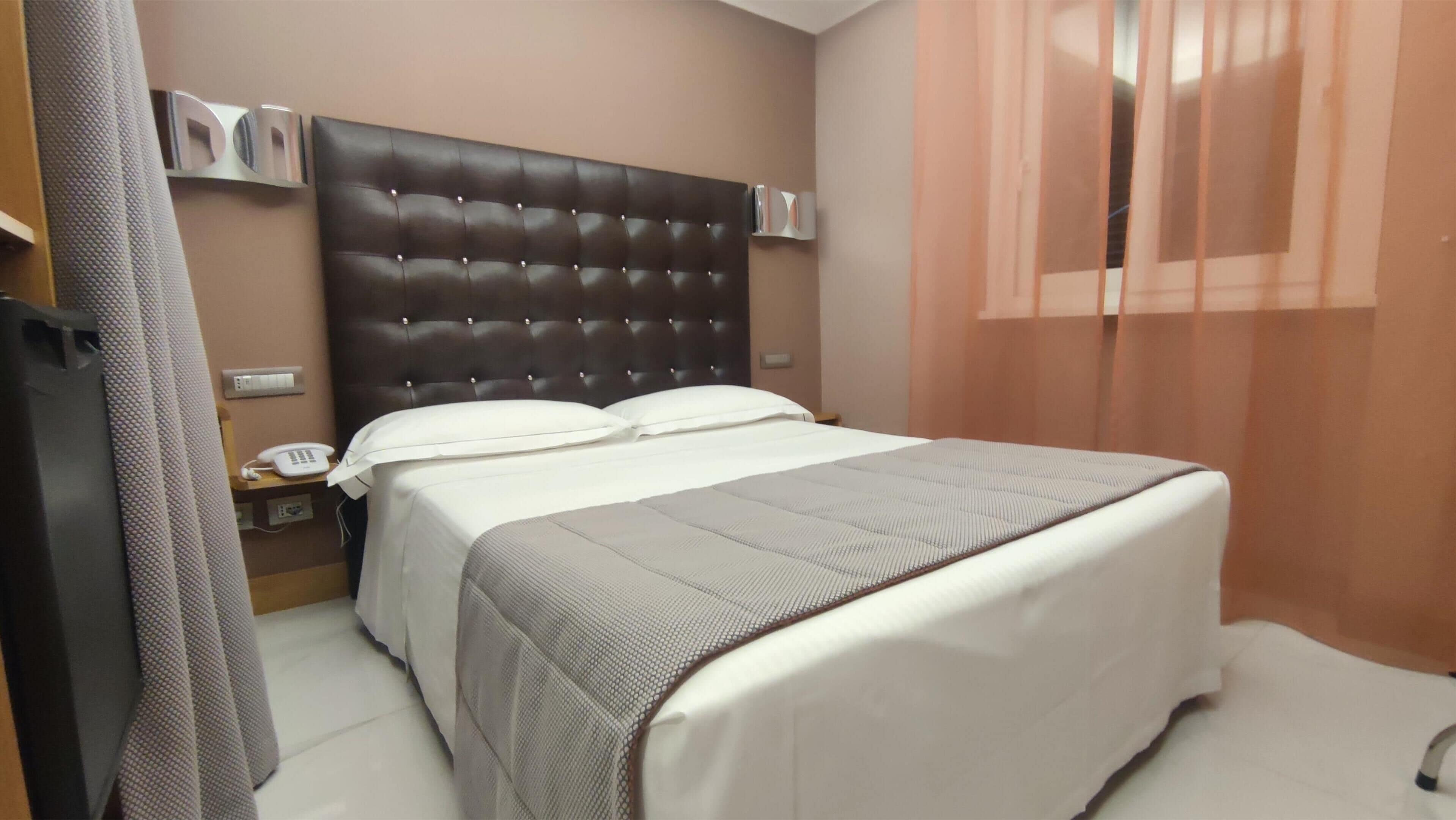 A neatly made bed with a tufted headboard, a bedside table with a phone, and a window with sheer curtains.