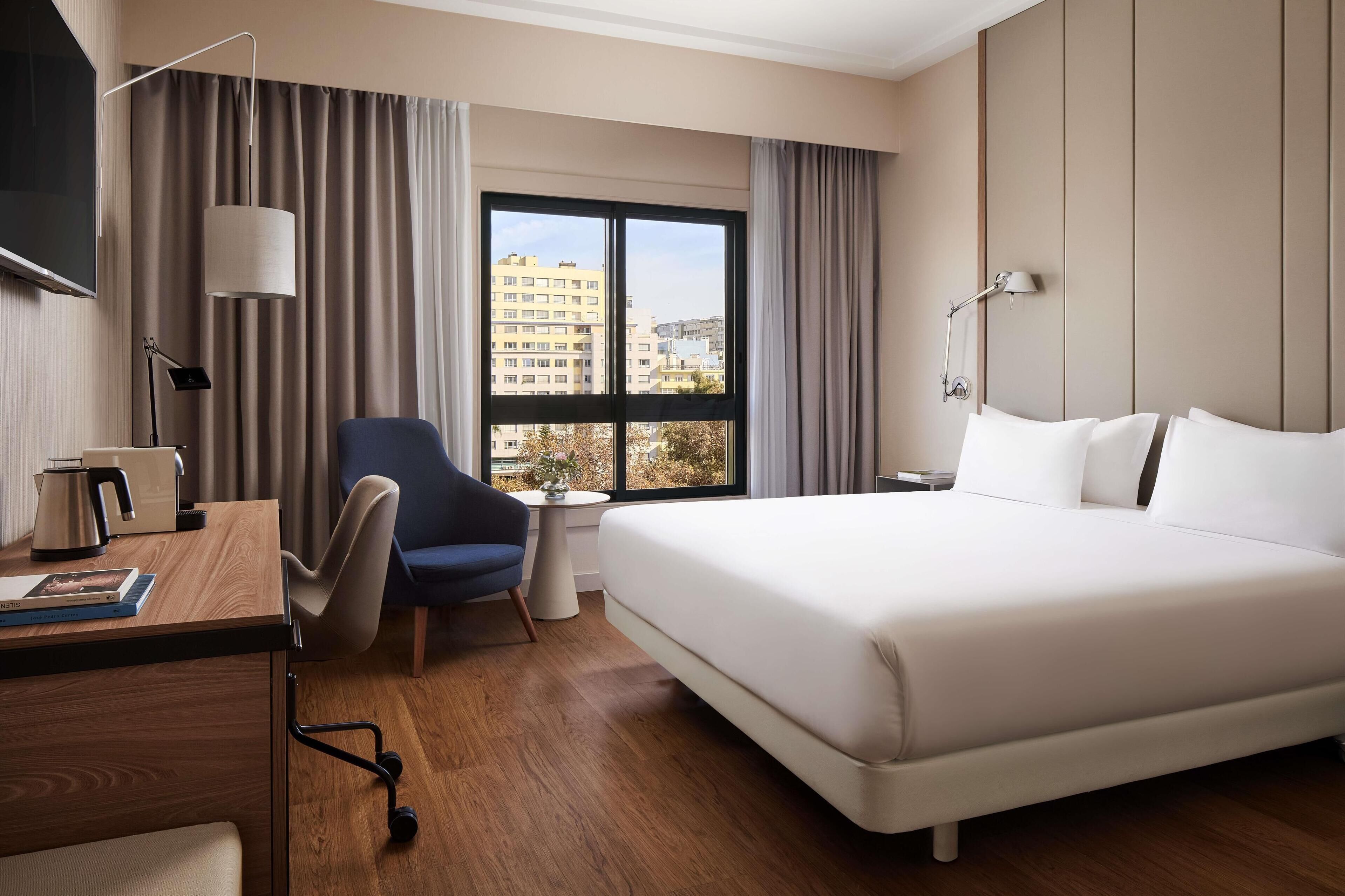 A hotel room with a large bed, a desk with a lamp, a chair, and a view of buildings through the window.