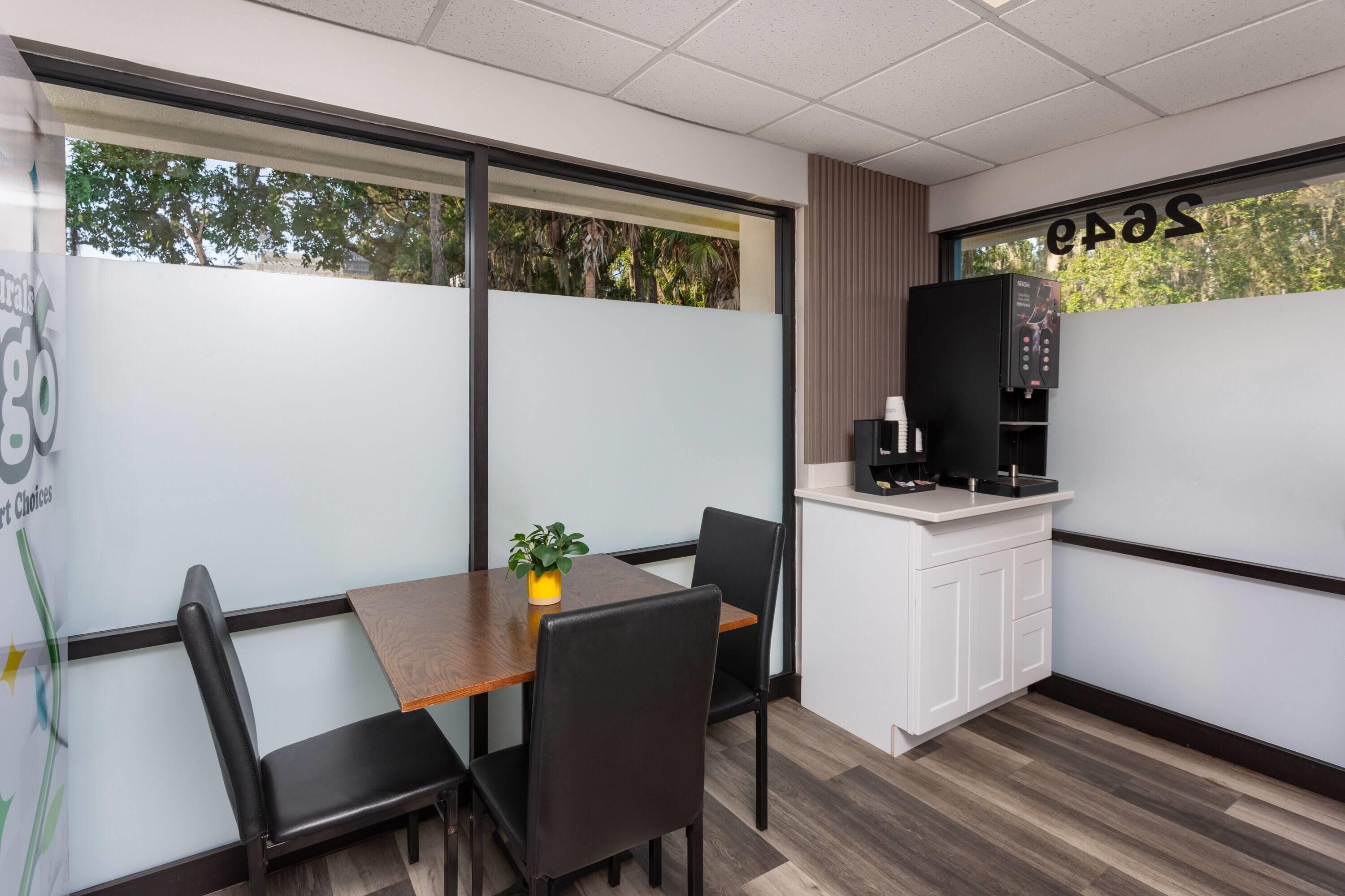 Una kitchenette pequeña y moderna con una cafetera, una mesa individual con dos sillas y una planta en maceta.