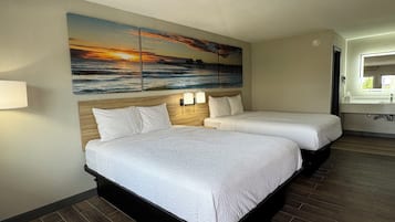 Une chambre d’hôtel avec deux lits, un tableau représentant un coucher de soleil sur l’océan, une tête de lit en bois, une table de chevet avec une lampe et une salle de bain avec un lavabo et un miroir.