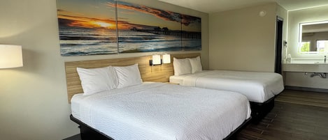 Une chambre d’hôtel avec deux lits, un tableau représentant un coucher de soleil sur l’océan, une tête de lit en bois, une table de chevet avec une lampe et une salle de bain avec un lavabo et un miroir.