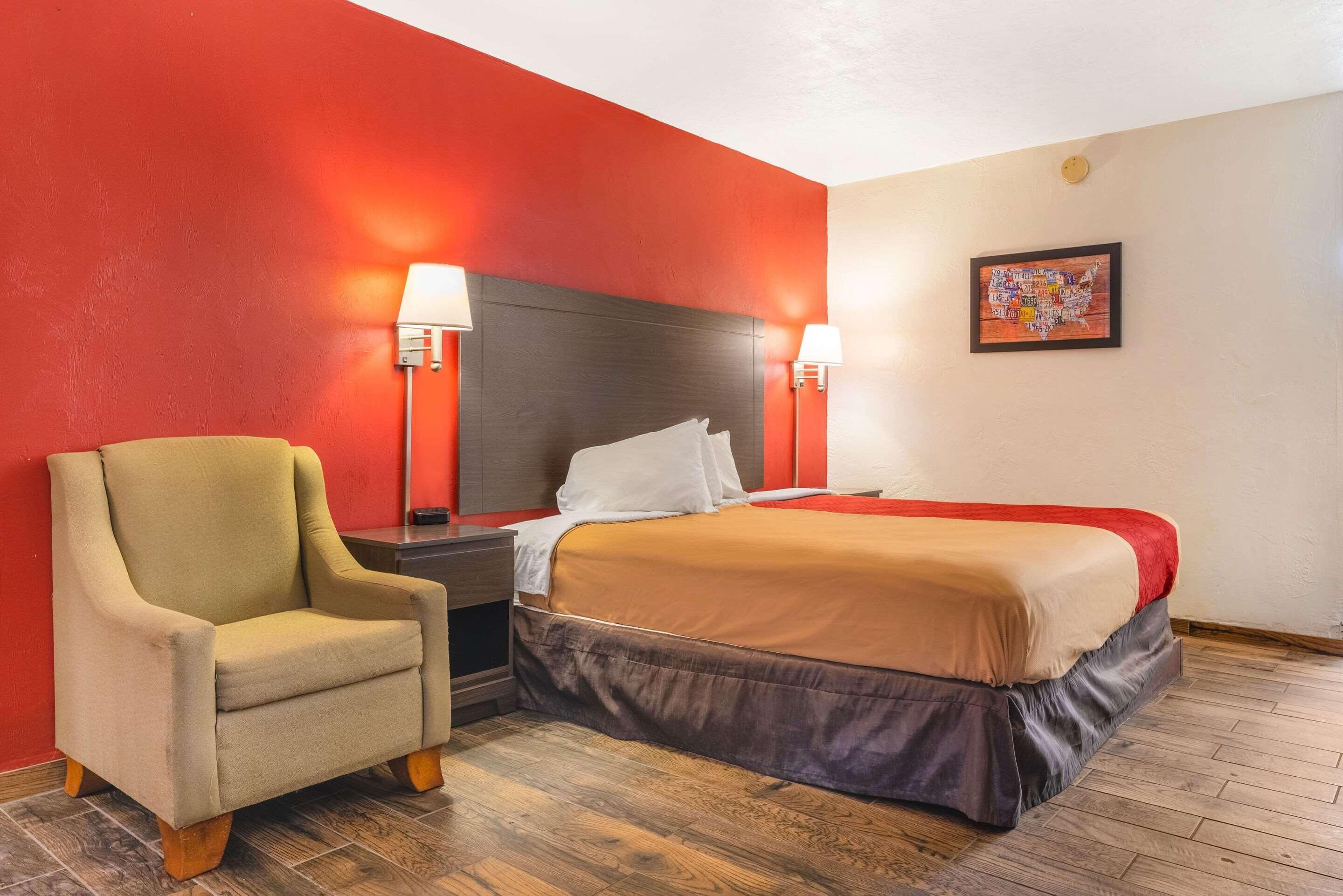 Una habitación de hotel con una cama, una silla, una mesita de noche y una lámpara fijada a la pared.