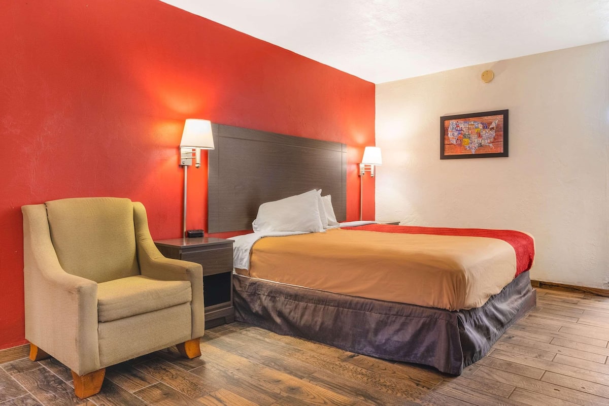 Una habitación de hotel con una cama, una silla, una mesita de noche y una lámpara fijada a la pared.