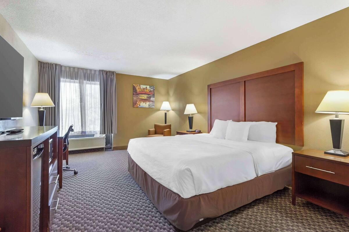 A hotel room with a large bed, two bedside tables with lamps, a desk with a chair, and a window with curtains.