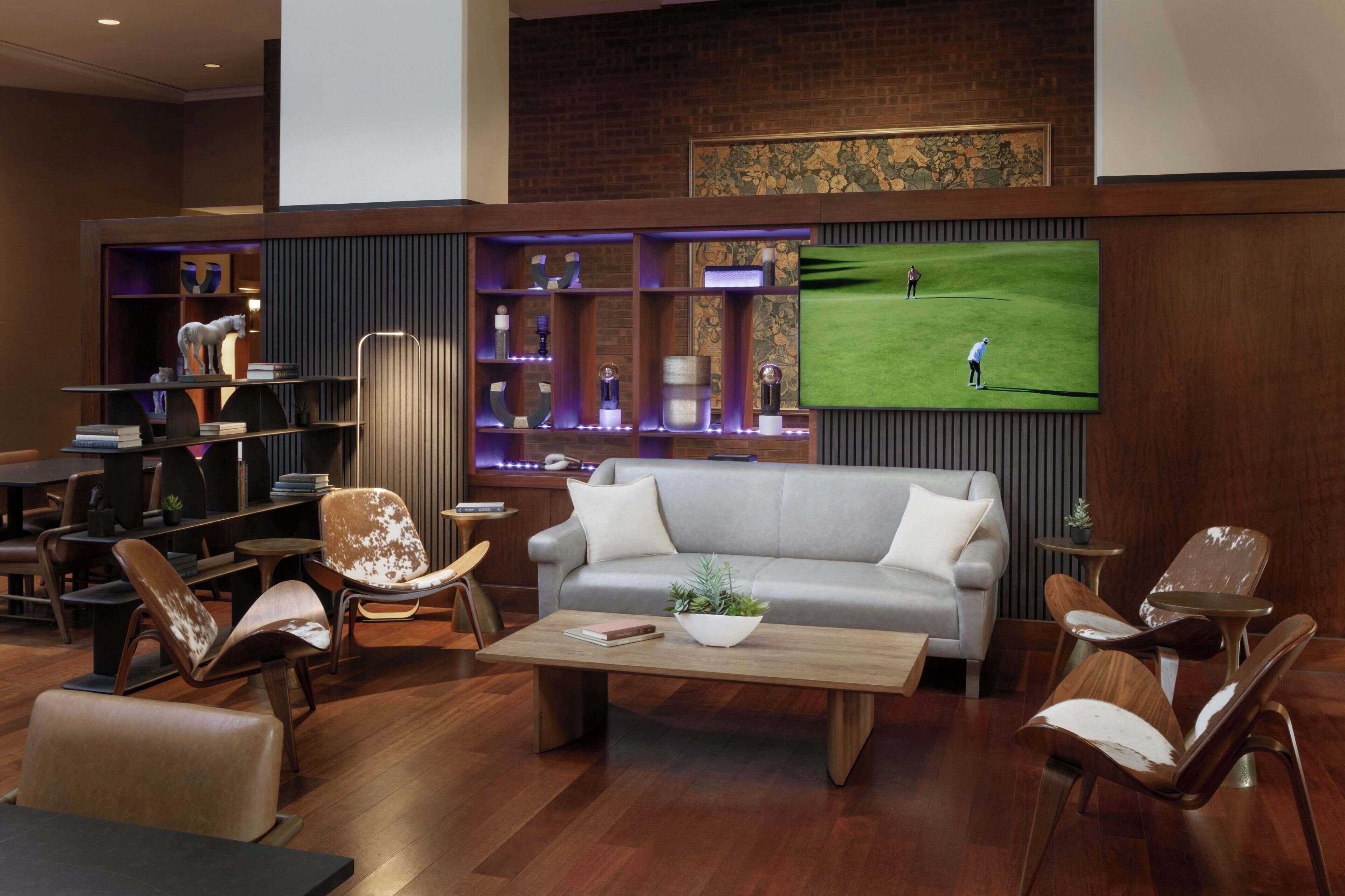 A modern lounge area with a grey sofa, leather armchairs, a wooden coffee table, and a TV displaying a golf game.