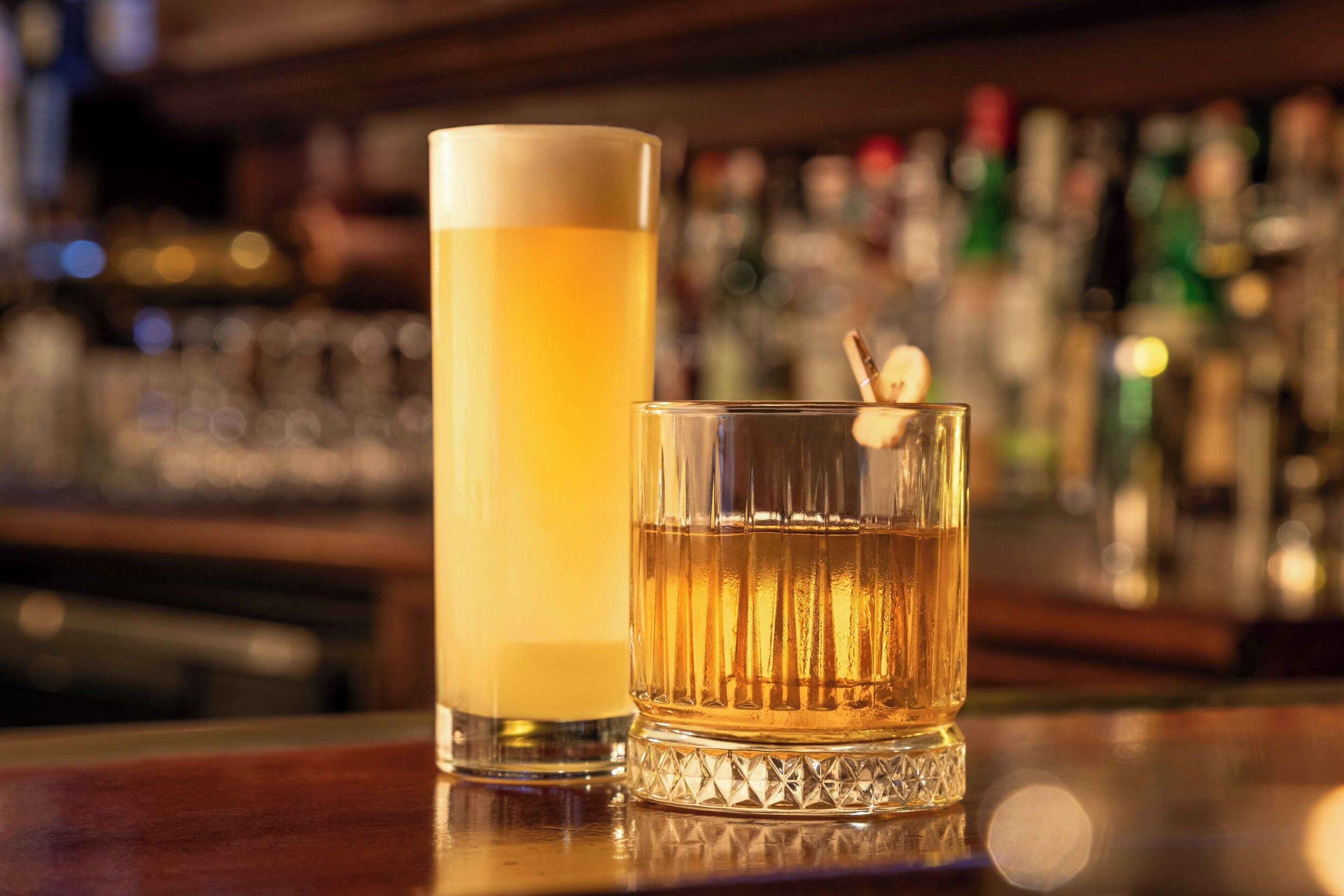 Two glasses of amber-colored beverages on a bar counter with a blurred background of bottles.