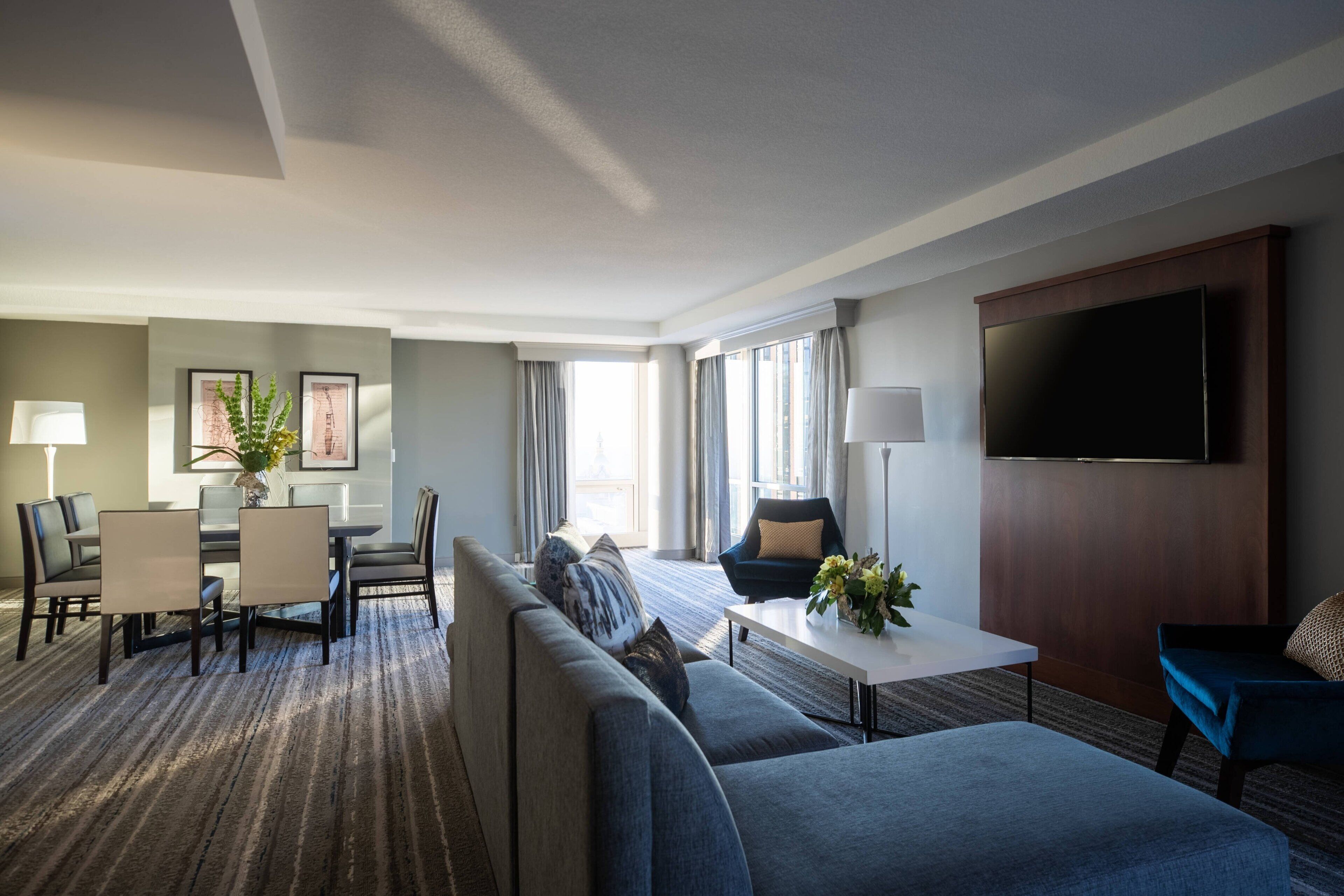 Habitación de hotel moderna con un sofá grande, una mesa de comedor con sillas, un televisor de pantalla plana en la pared y un ventanal que proporciona luz natural.