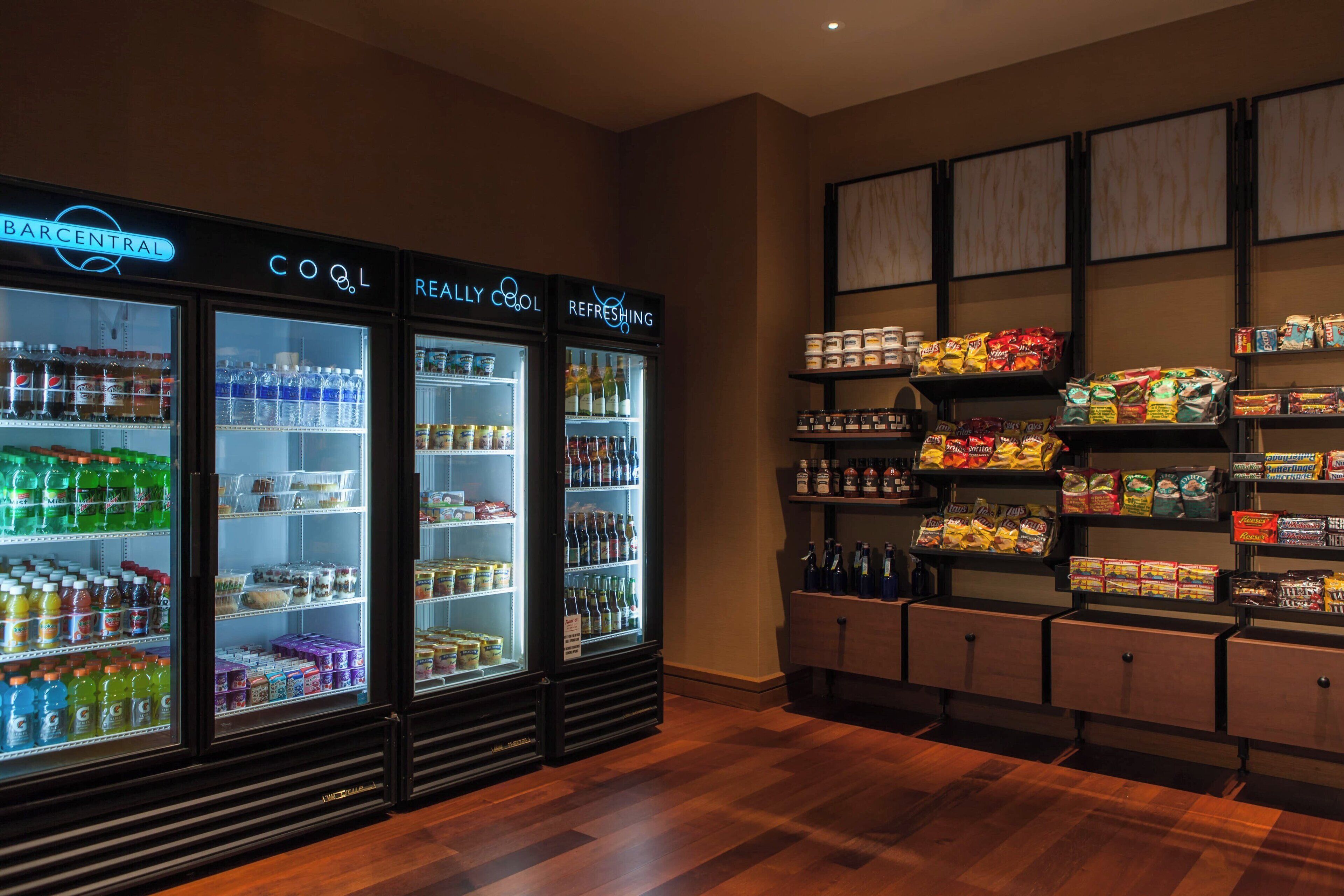 Een winkel met drie gekoelde drankvitrines, gelabeld 'BARCENTRAL', 'COOL' en 'REFRESHING'. Planken zijn gevuld met snacks en dranken.
