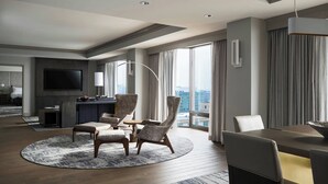 Une chambre d’hôtel moderne dotée d’un grand téléviseur à écran plat, d’un coin repas avec une table ronde et des chaises, d’un canapé et offrant une vue sur la ville grâce à de grandes fenêtres.