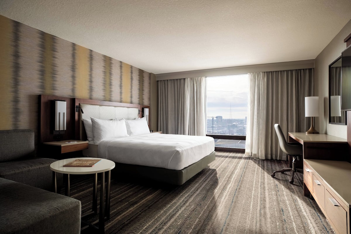 Hotelkamer met een groot bed, een bureau, een stoel, een kleine ronde tafel en uitzicht op de stad door grote ramen.