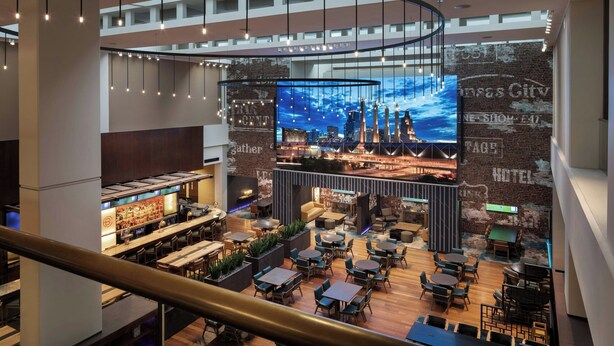 Un hall d’hôtel moderne doté d’un bar, de différents espaces de détente et d’un grand écran numérique affichant une vue panoramique sur la ville.