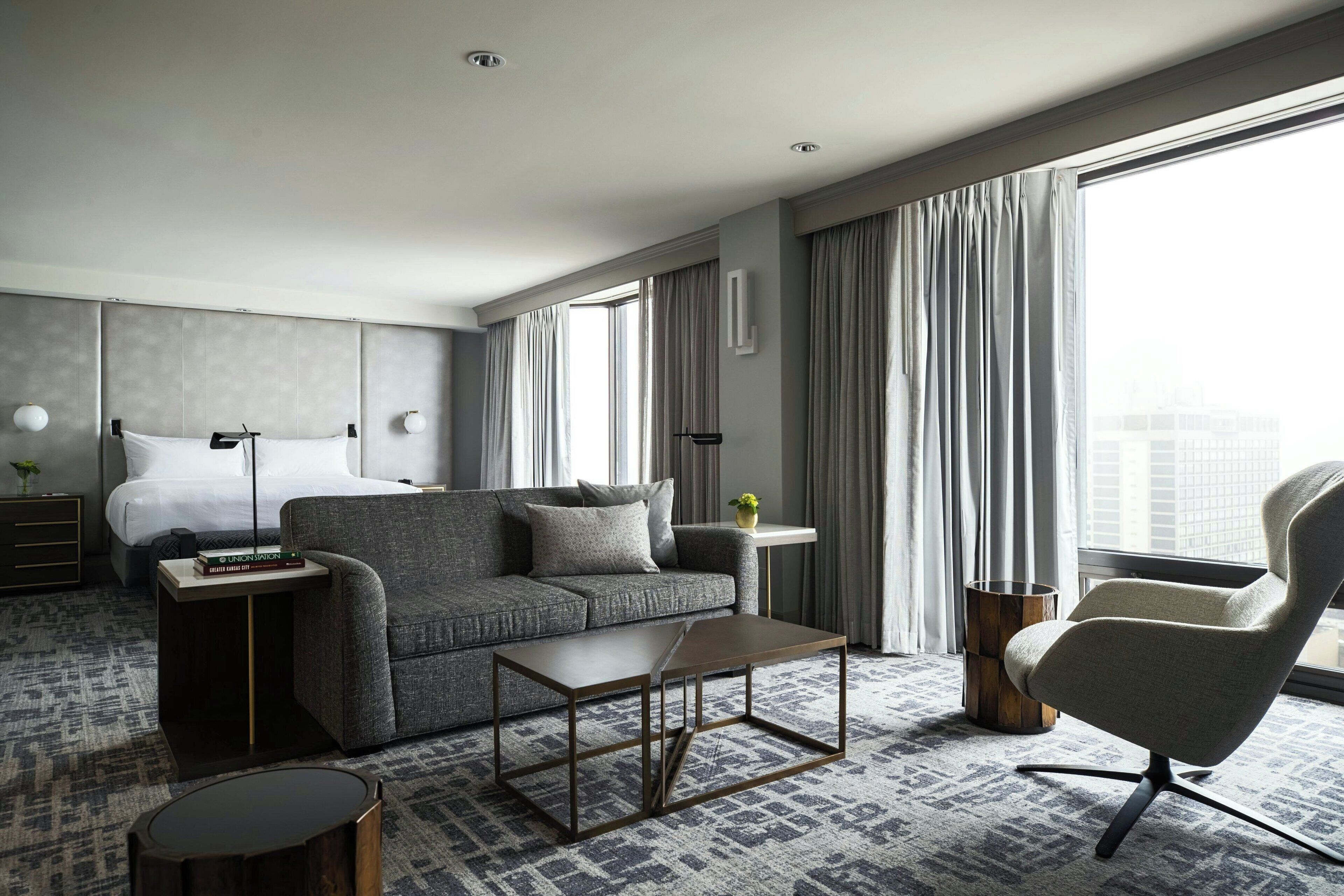 Una habitación de hotel moderna con un sofá gris, una pequeña mesa de centro, una silla, una mesita de noche con lámpara y un amplio ventanal con cortinas.