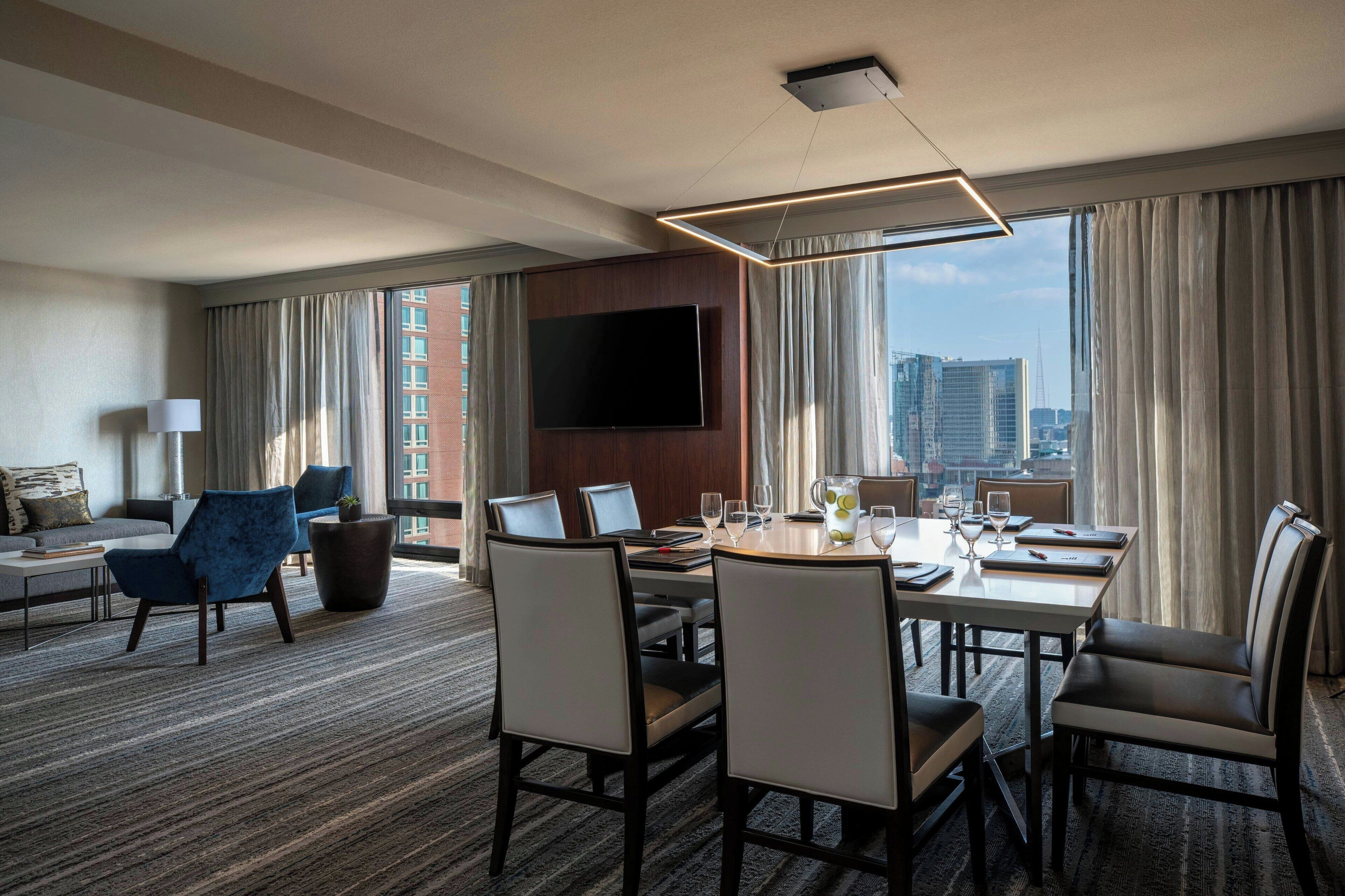 A hotel room with a dining area, a sofa, a TV, and a view of the city.