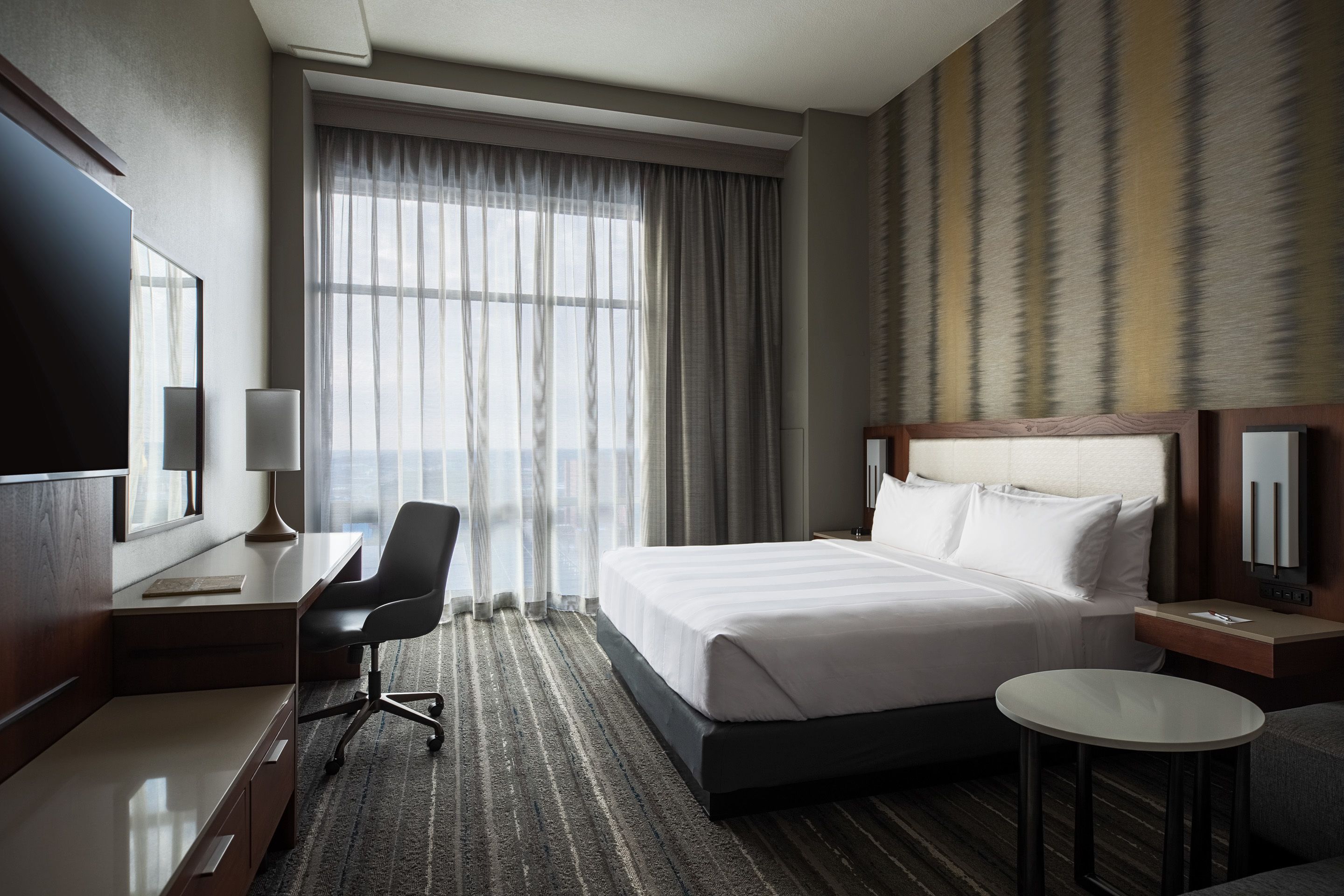 Habitación de hotel con una cama grande, un escritorio con silla, una mesita redonda y un televisor.